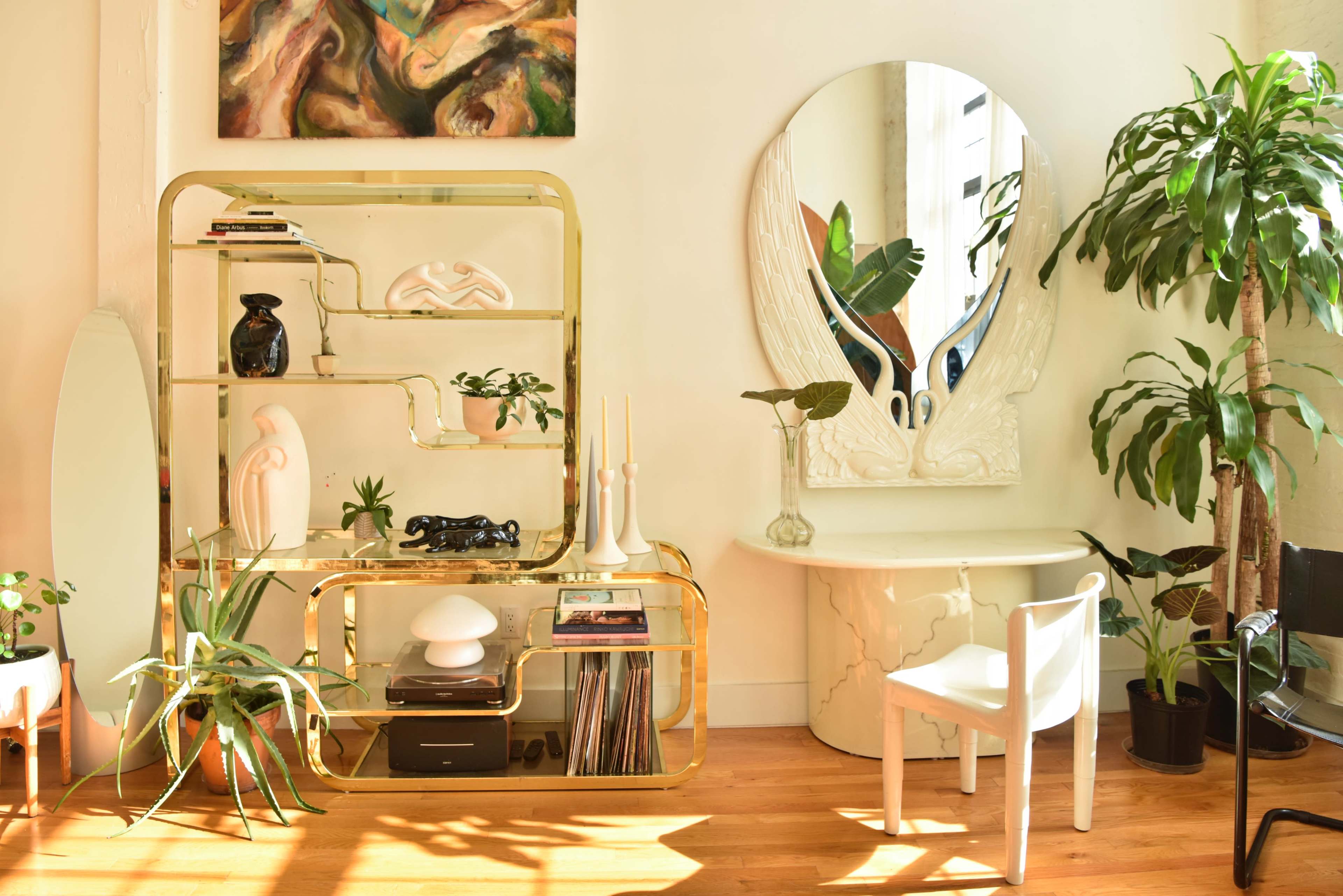 A bright interior scene features a gold shelving unit, a decorative mirror, a small table, potted plants, and various decorative objects arranged in a sunlit space.