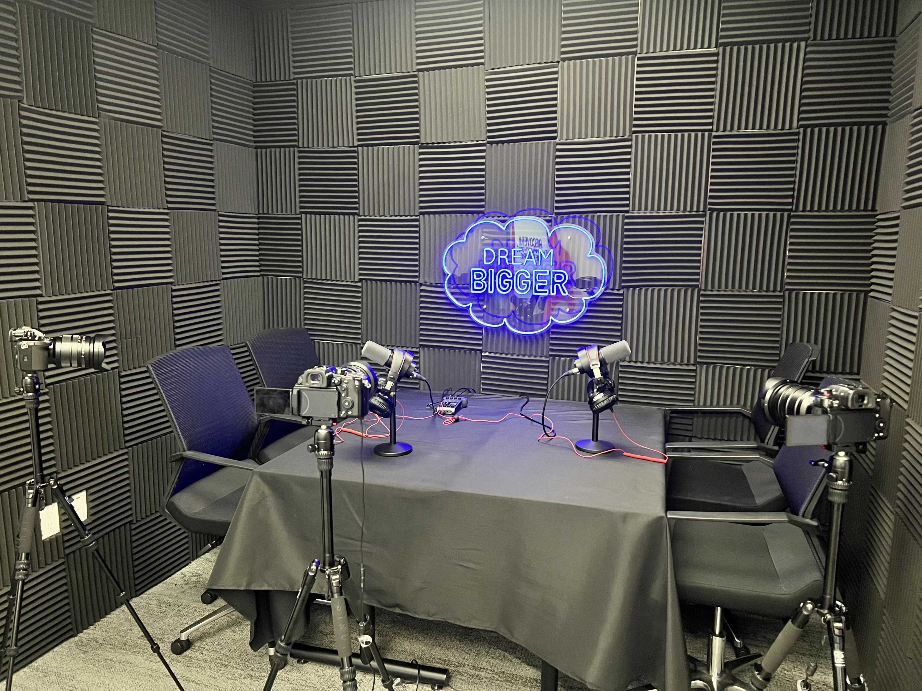 A podcast recording studio with four chairs, microphones, and cameras set up around a table, against a soundproof wall with a blue neon sign that reads "Dream Bigger."