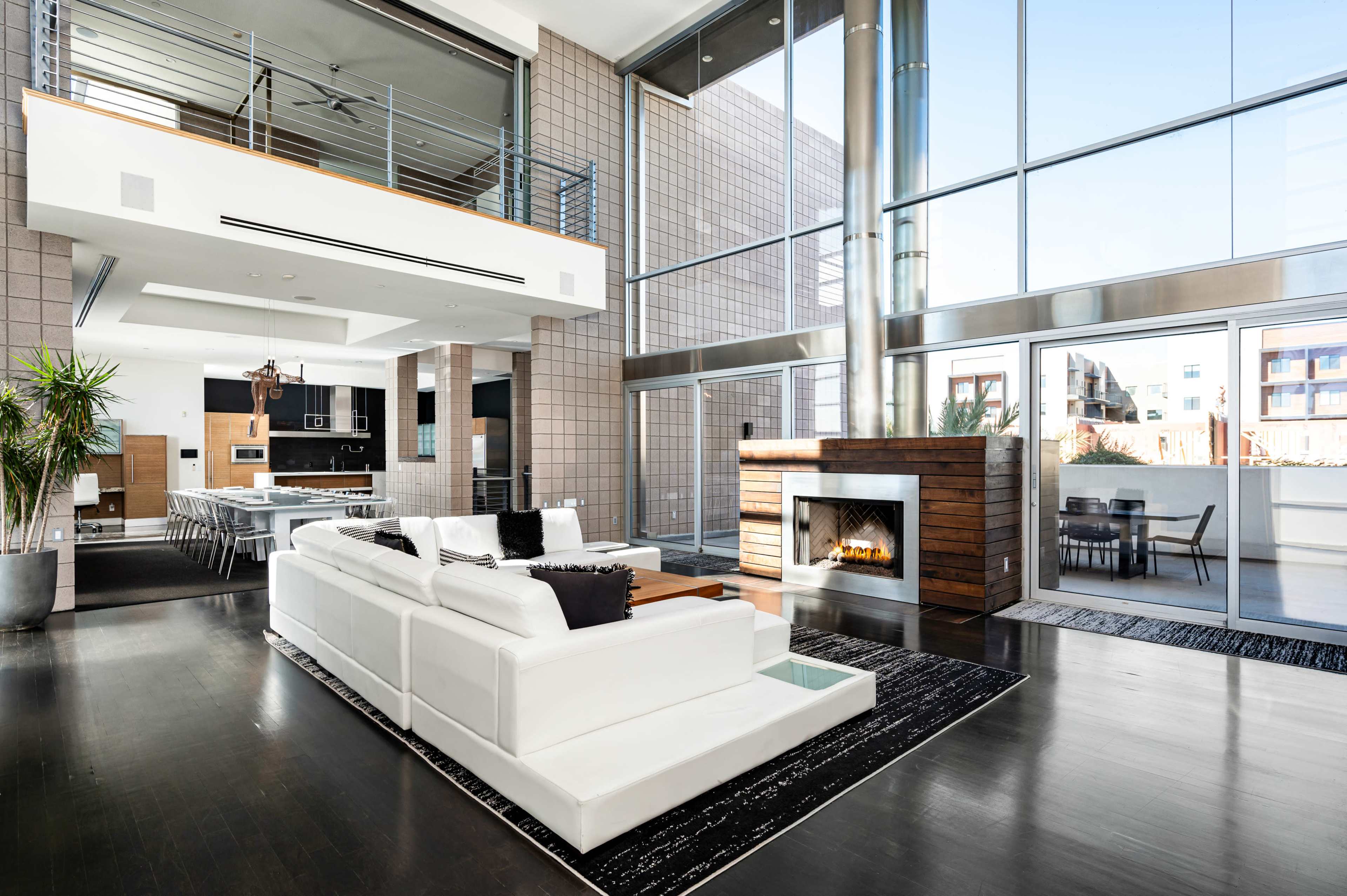 A spacious modern living room features a large white sectional sofa, a fireplace, and floor-to-ceiling windows that provide ample natural light.