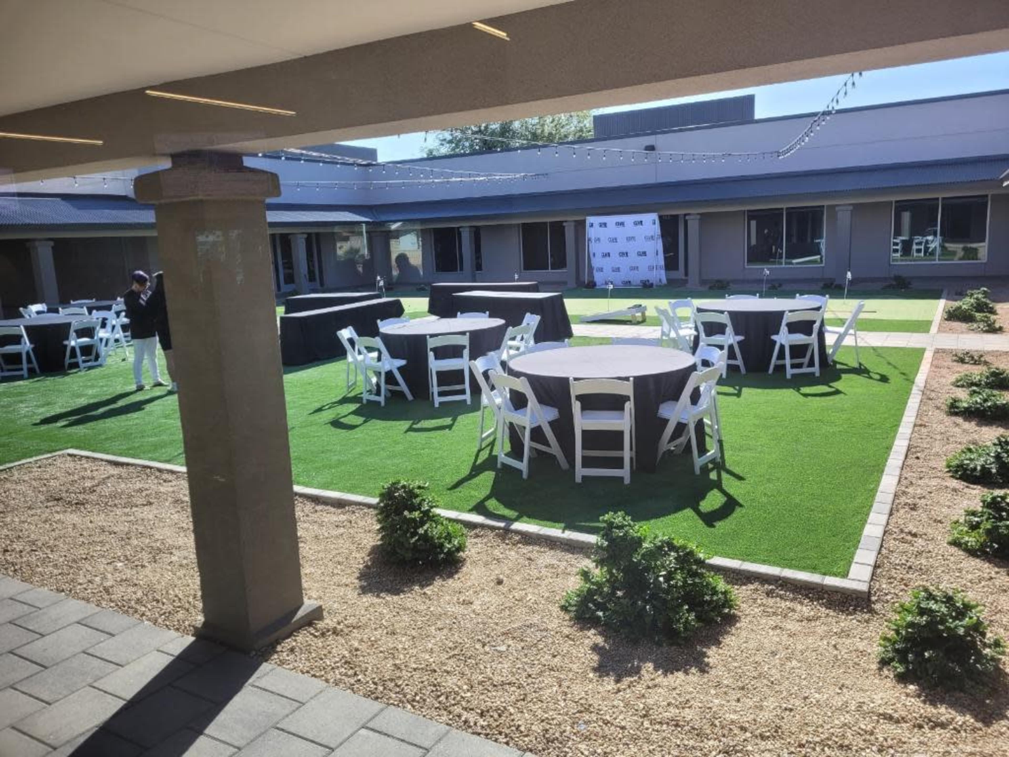 An outdoor event space with several round tables set up on grass and surrounded by hedges.