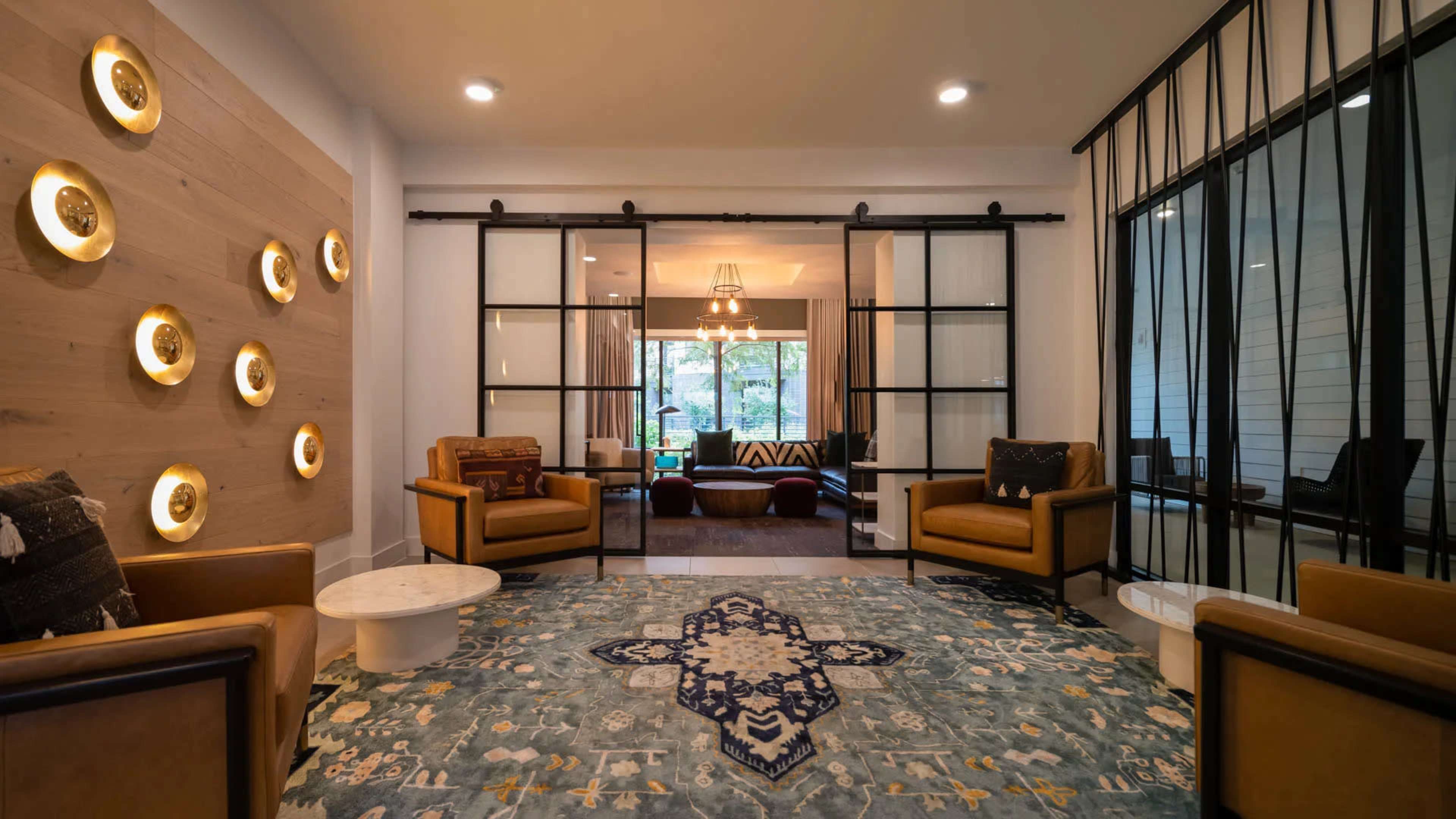 The image shows a modern lounge area with two brown armchairs, a decorative rug, and sliding glass doors leading to a well-lit interior space.