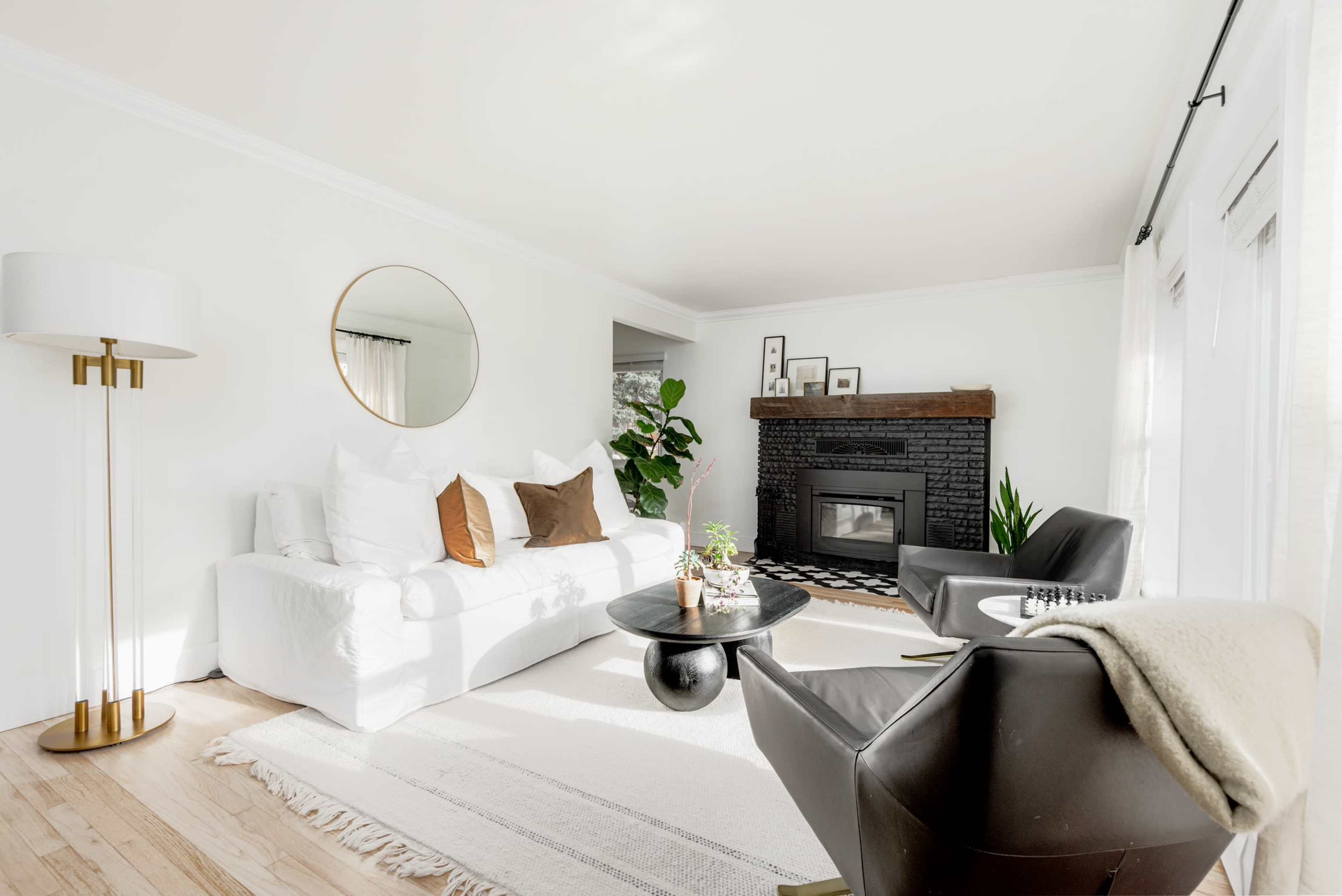 The living room features a white sofa, a black accent chair, a round coffee table, and a fireplace with a dark mantel, all set against light walls and wooden flooring.