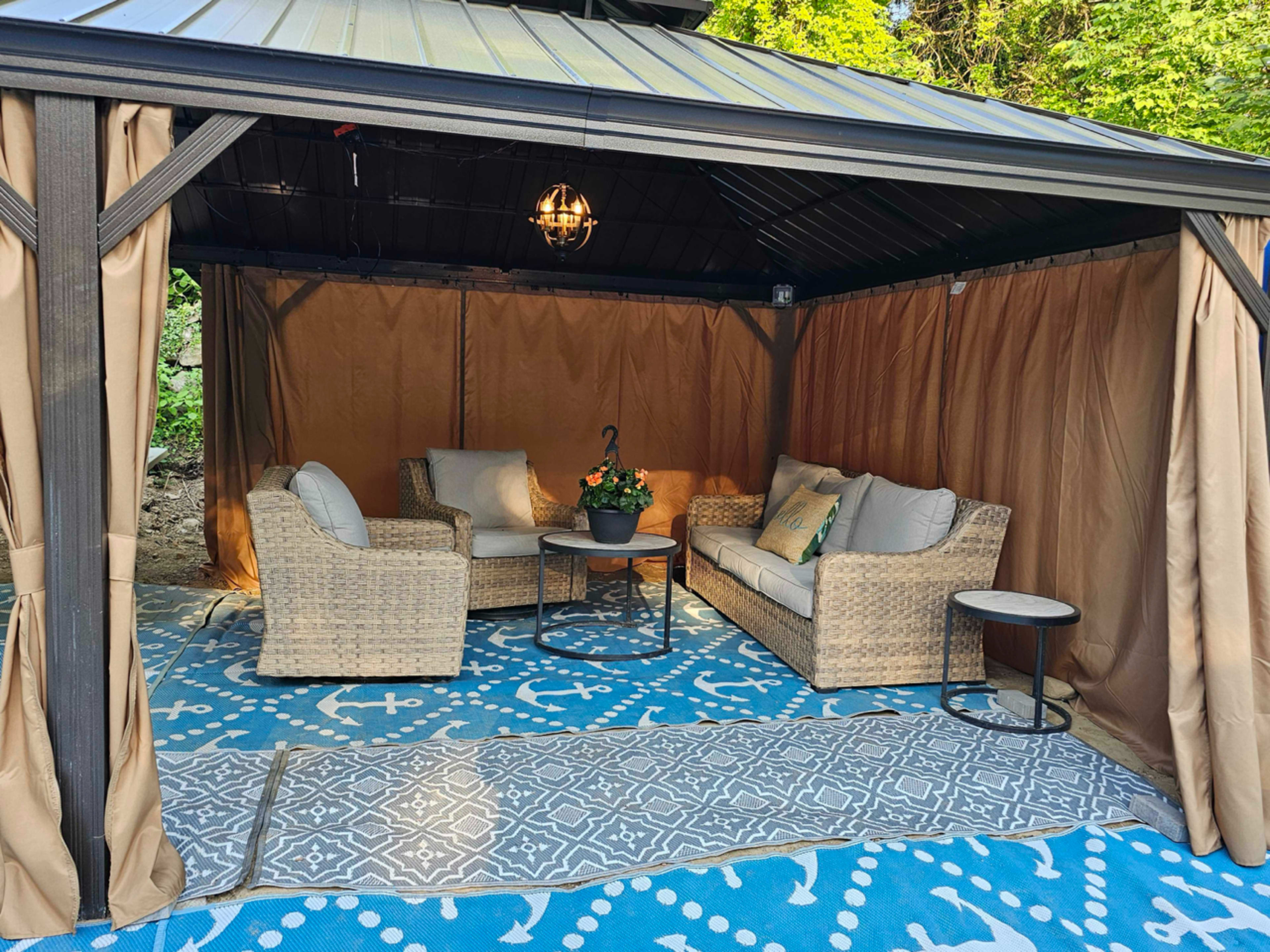 A cozy outdoor gazebo features a seating arrangement with a sofa, two chairs, and a side table on a patterned rug, surrounded by draped curtains.