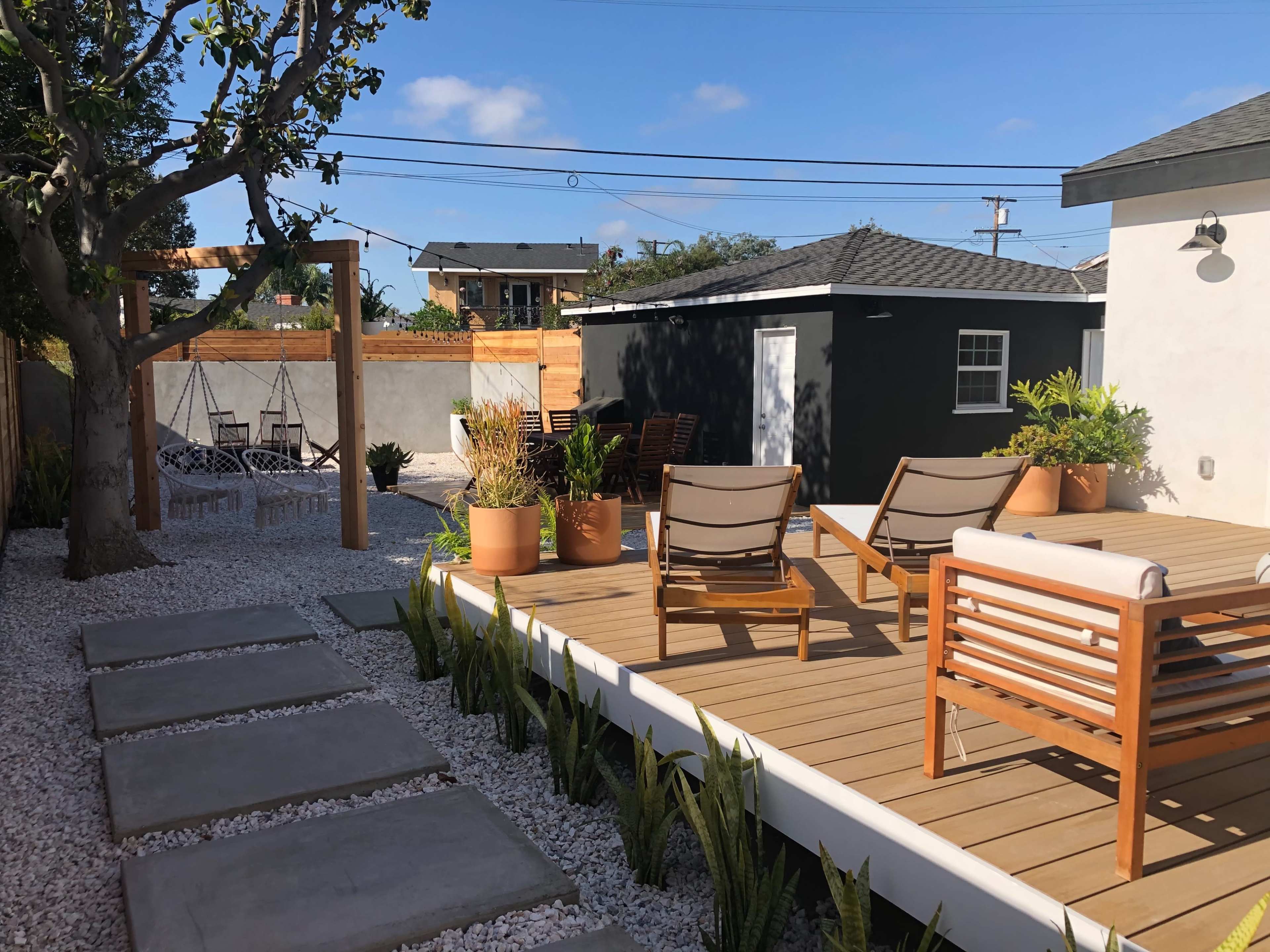 A landscaped backyard with a wooden deck, lounge chairs, and stone pathways leading to a seating area surrounded by potted plants and gravel.