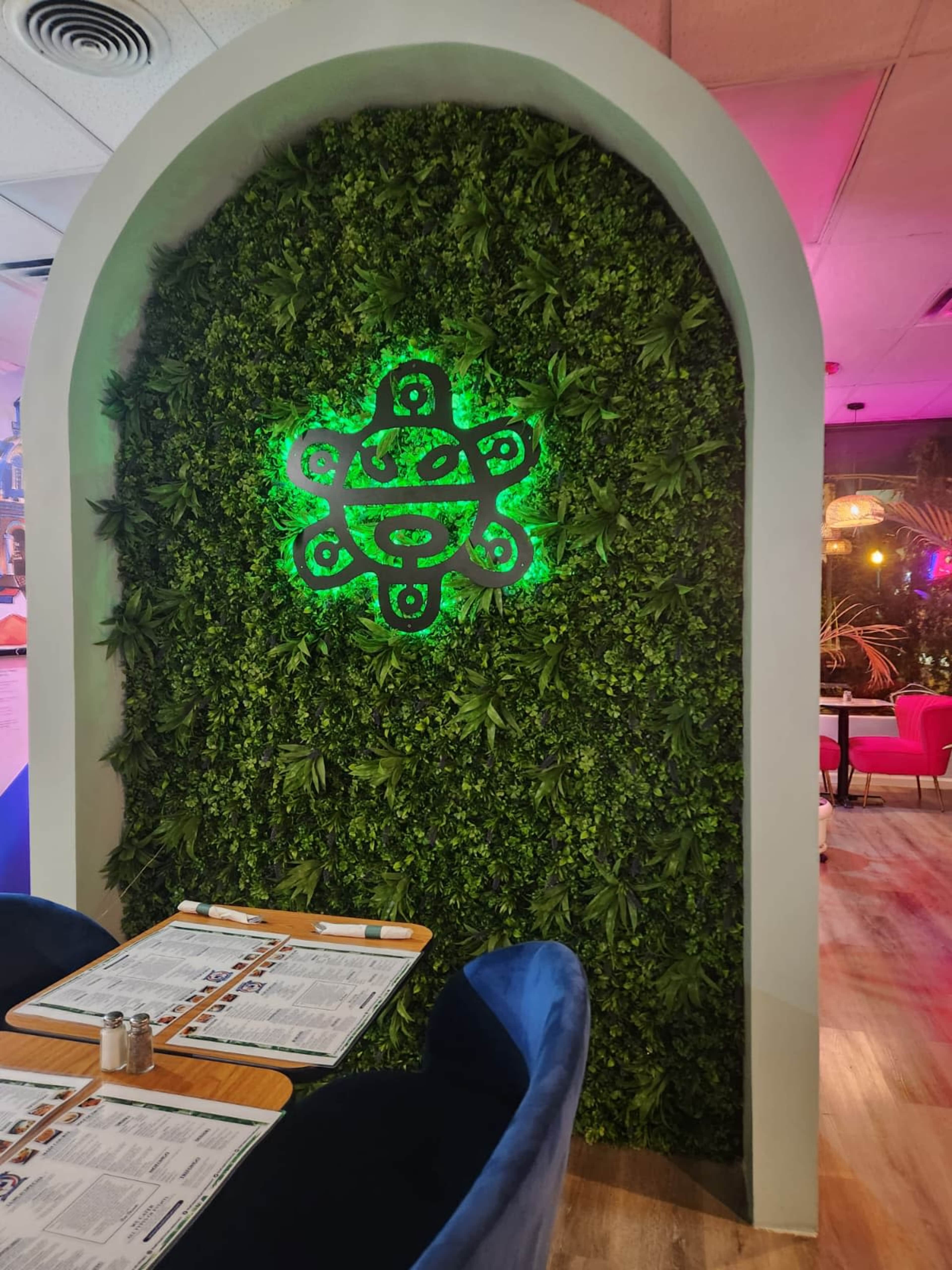 The image shows a green wall decorated with artificial plants and a glowing logo, set within a light-colored archway, with wooden tables and menus in the foreground.