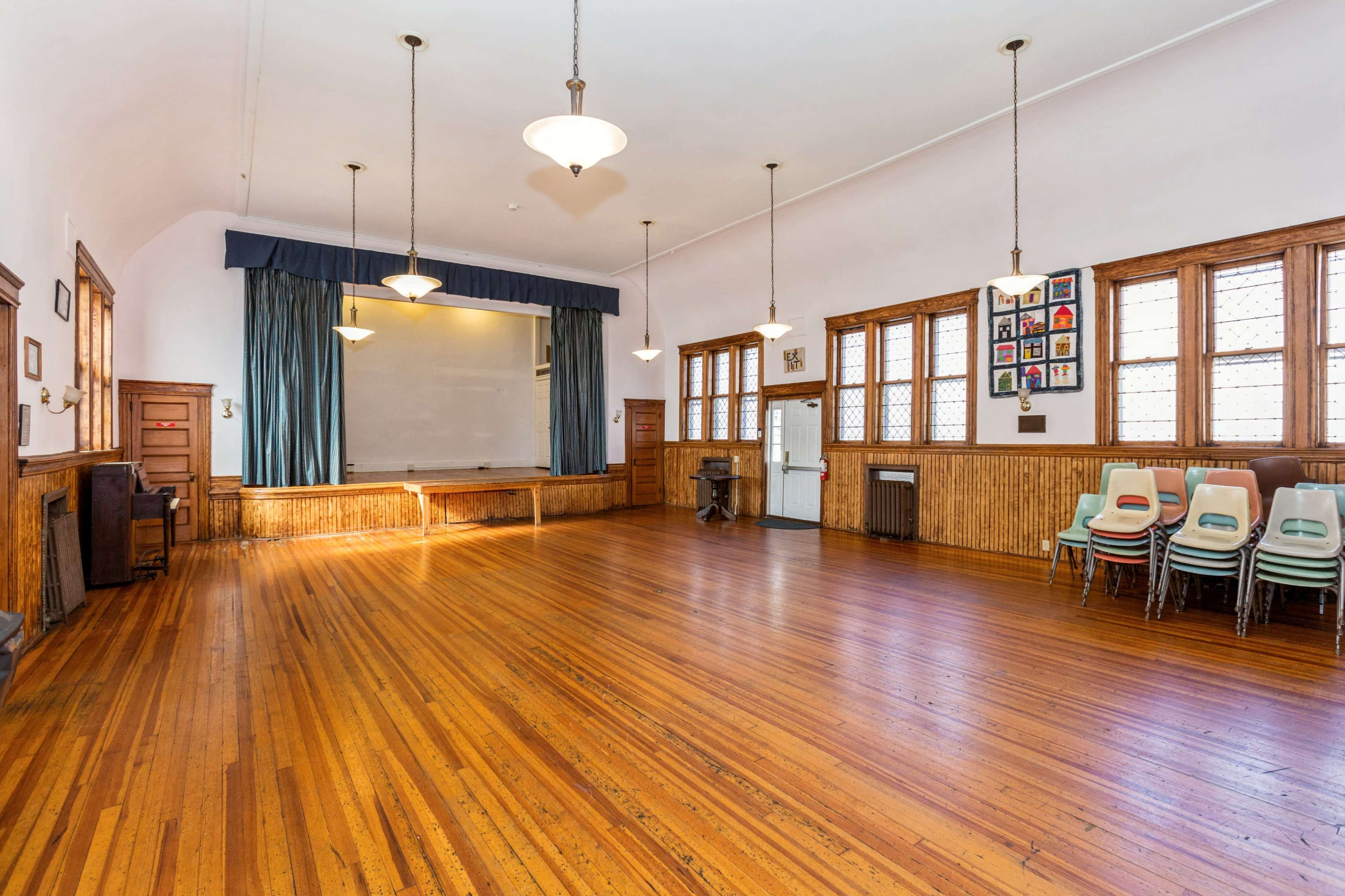 An empty wooden-floored hall with a stage at one end, large windows along the walls, and stacked chairs in one corner.