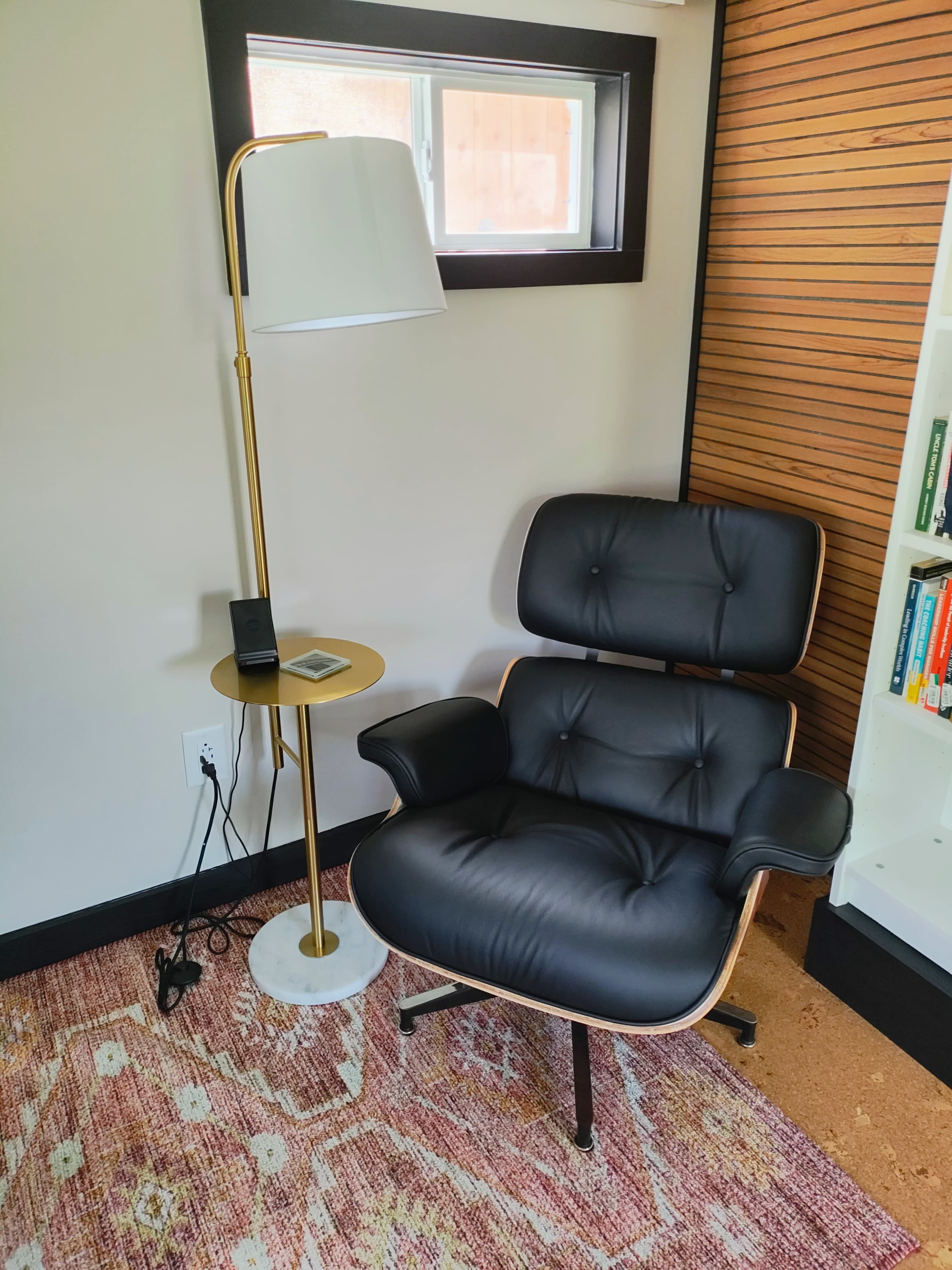 The image shows a modern black chair next to a gold floor lamp and a small side table with a device on it, set against a light-colored wall and a wooden bookshelf.