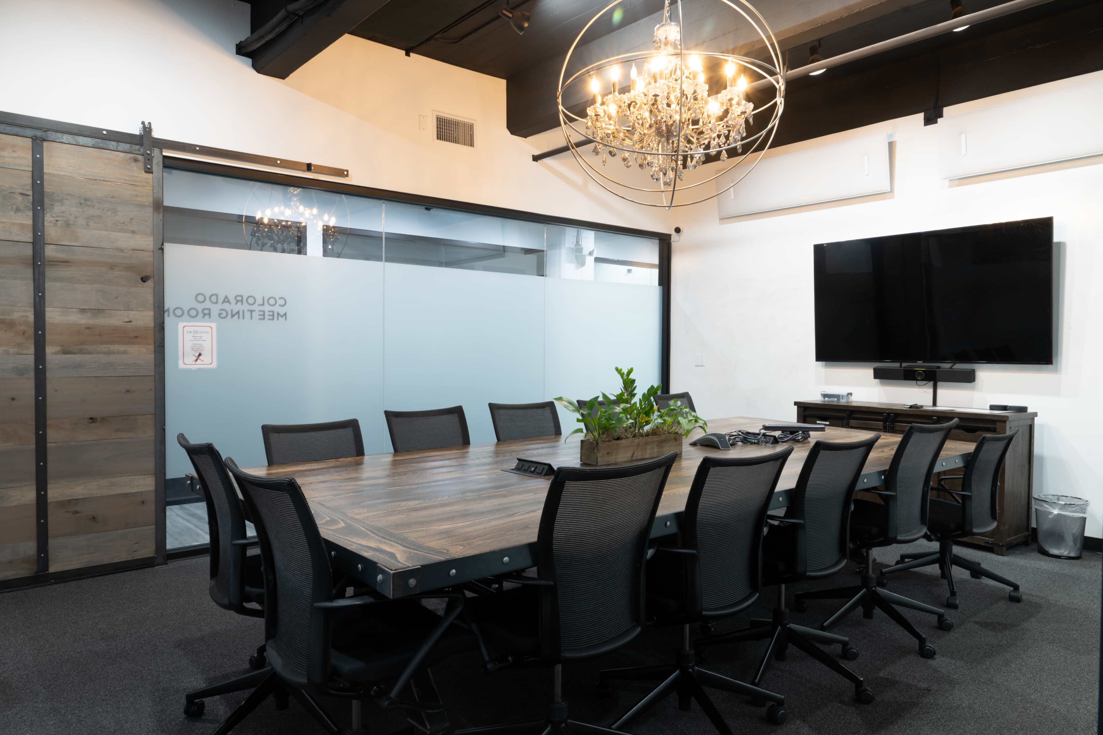 The image shows a modern conference room with a large wooden table surrounded by black office chairs, a TV mounted on the wall, and a glass partition with a plant centerpiece.