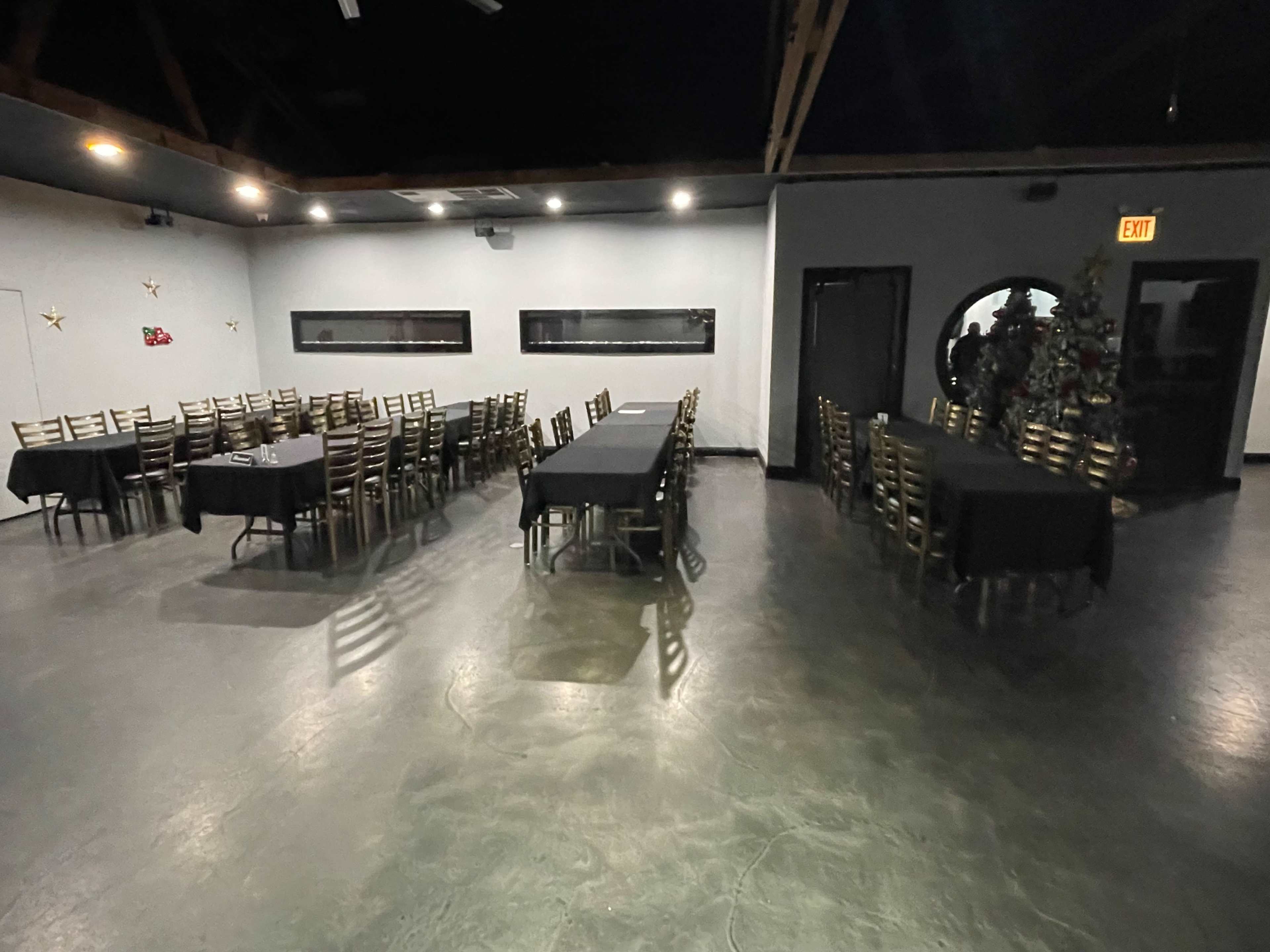 A spacious dining area with multiple long tables covered in black tablecloths arranged in rows, surrounded by metal chairs.