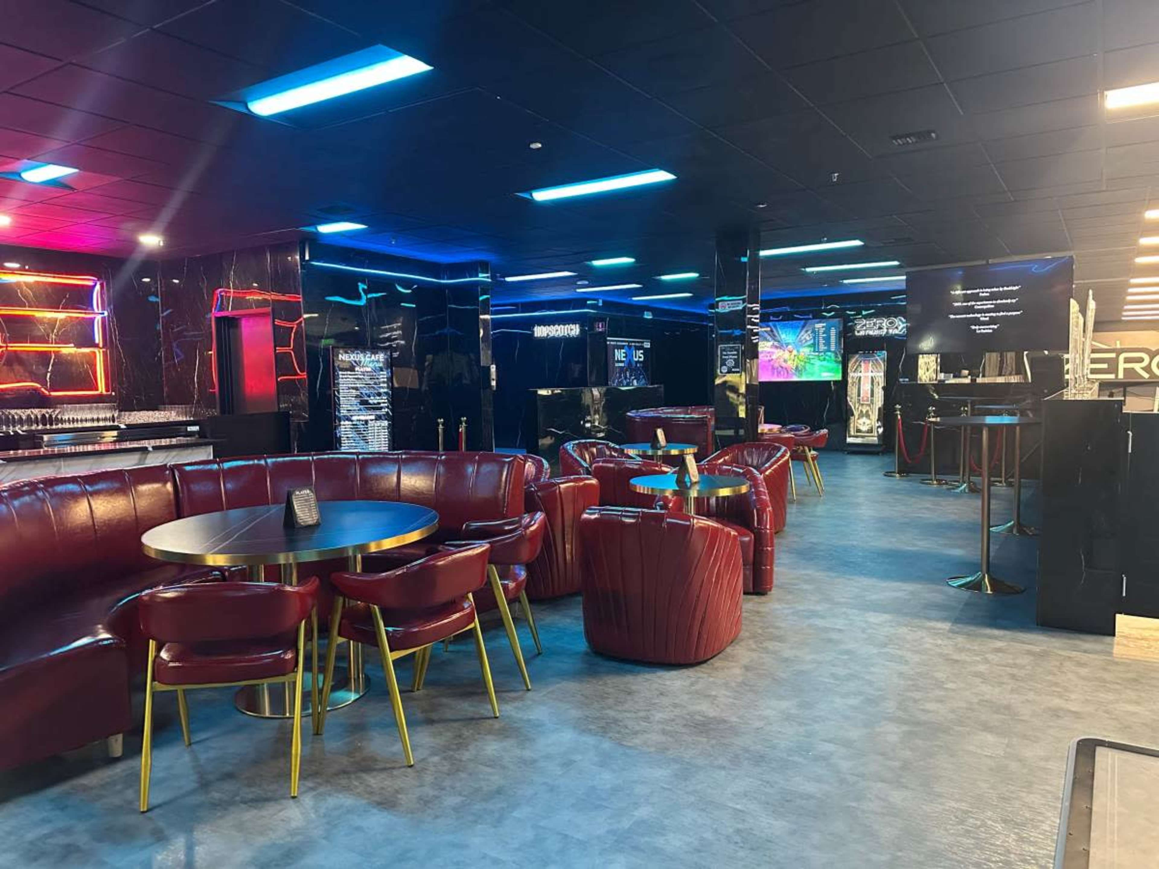 The image shows a modern lounge area featuring red leather seating, round tables, and illuminated walls in a dimly lit environment.