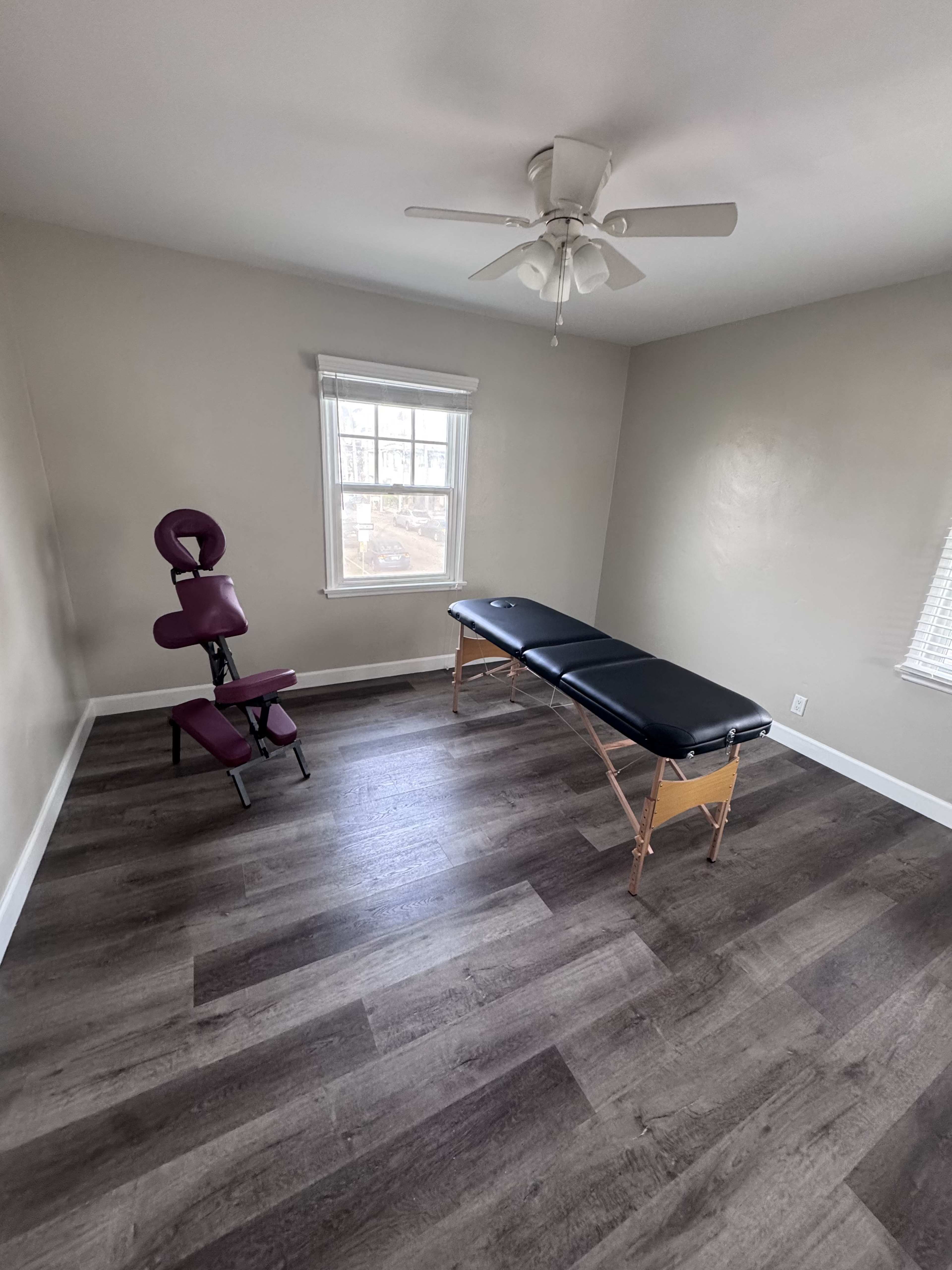The image shows a sparsely furnished room with a massage table and an ergonomic chair, along with a ceiling fan and windows letting in natural light.