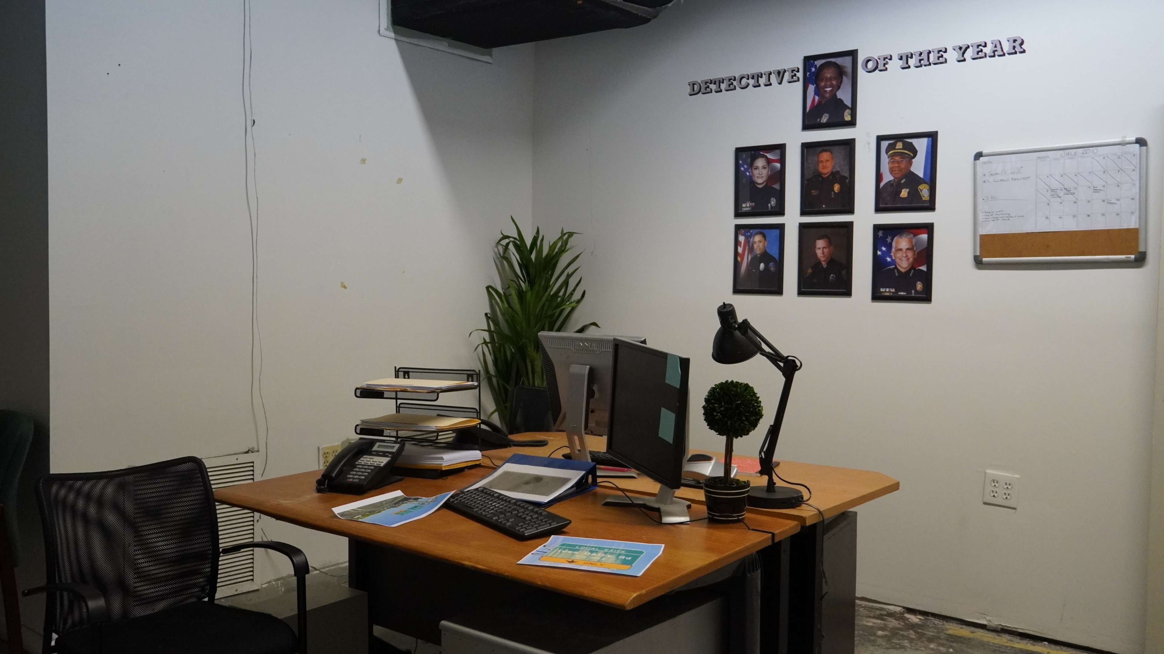 The image depicts an office with a wooden desk, an office chair, two computer monitors, a phone, and framed photographs of individuals on the wall above the desk, labeled "DETECTIVE OF THE YEAR."