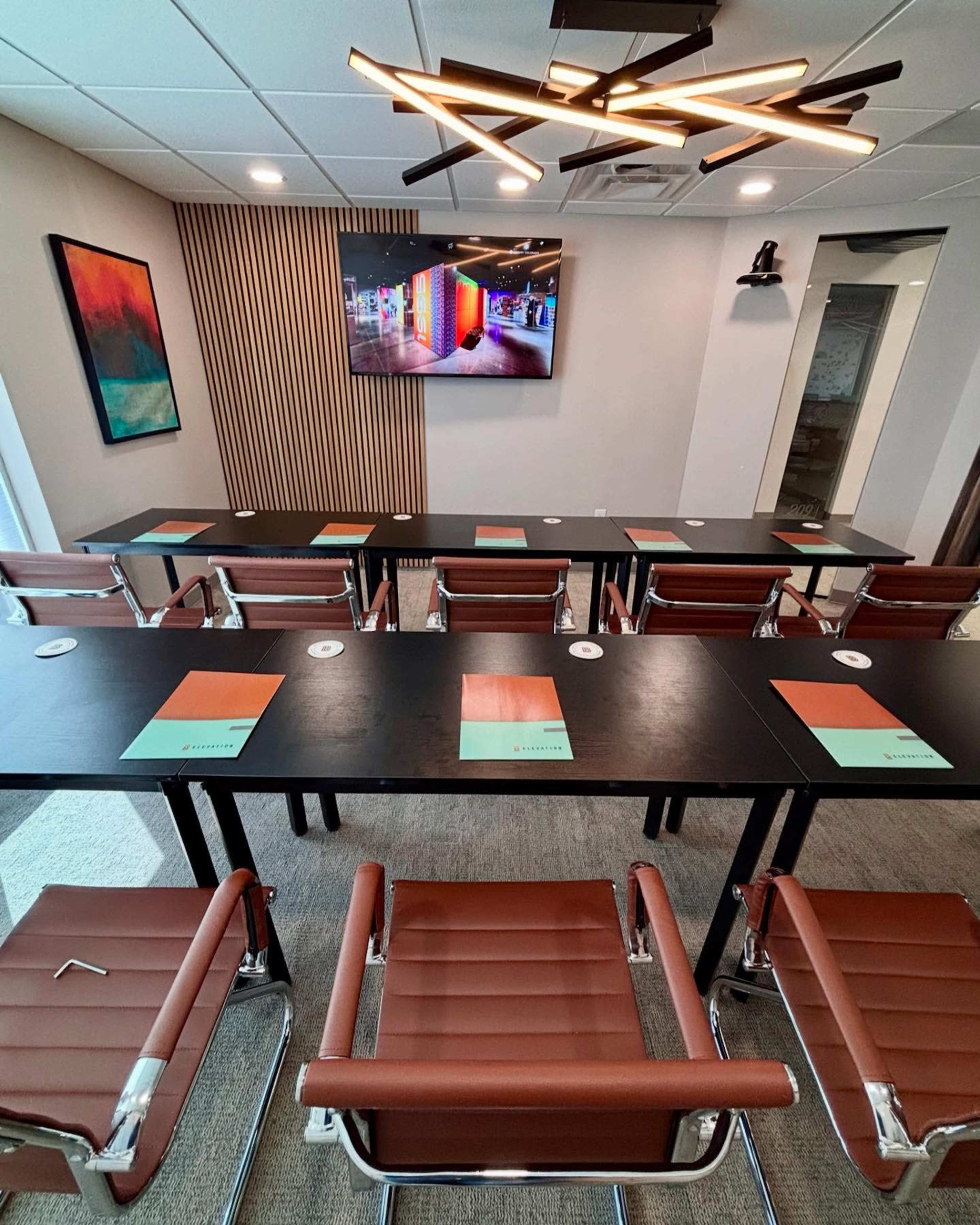 The image shows a modern conference room with lined tables, brown leather chairs, a television displaying colorful graphics, and contemporary lighting fixtures.