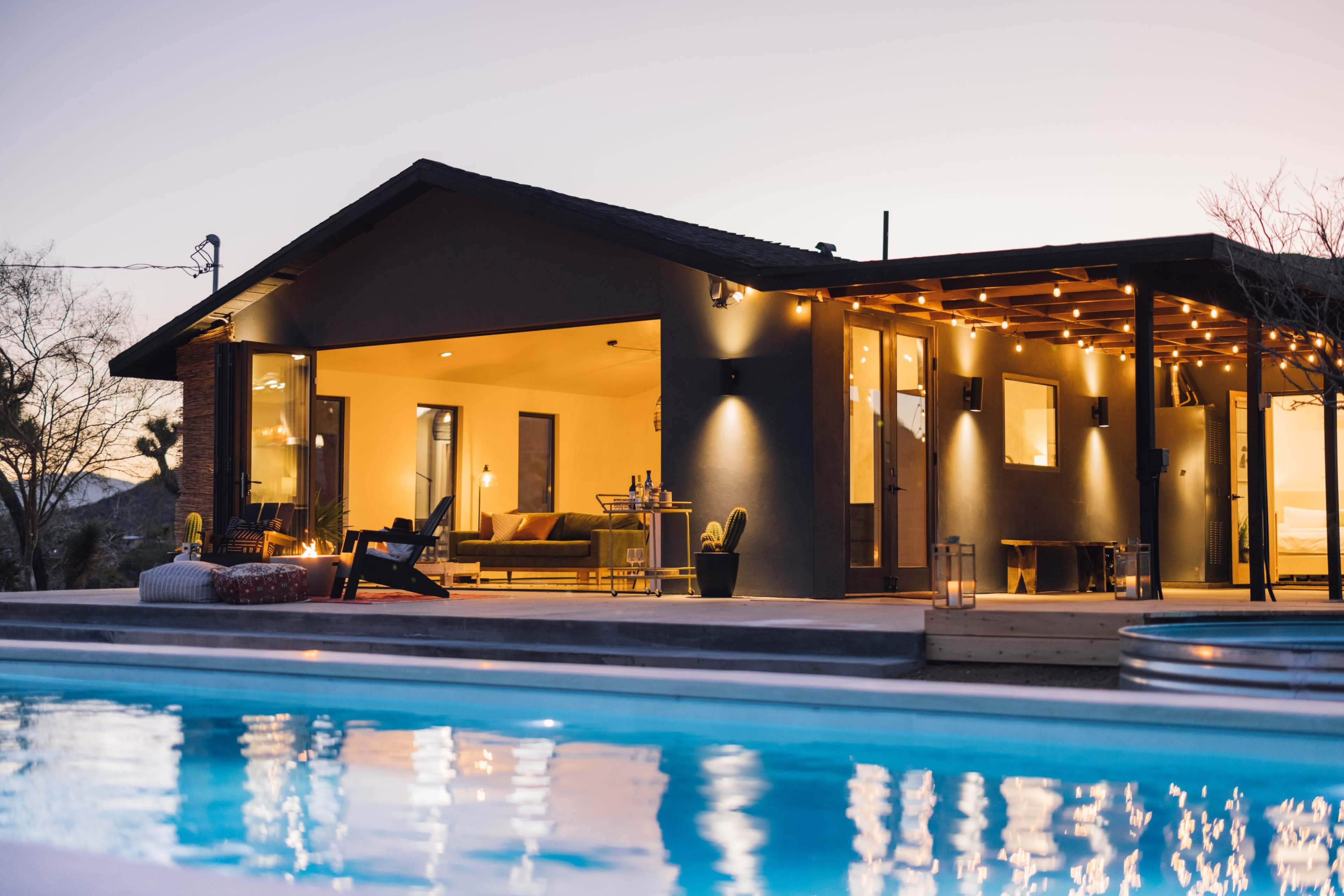 A modern house with large windows and outdoor seating overlooks a pool at dusk.