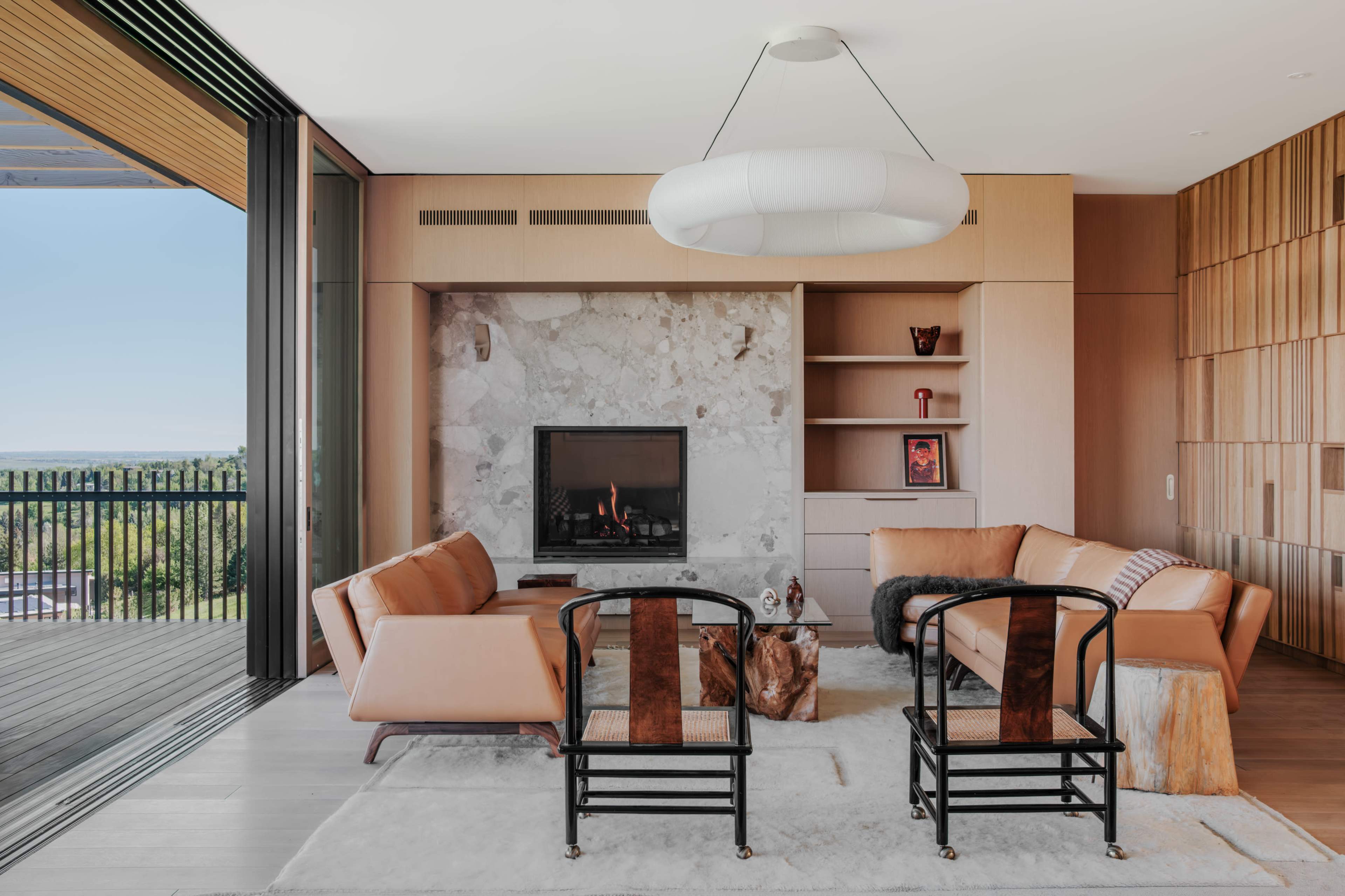 The image shows a modern living room featuring a marble fireplace, two leather sofas, and black wooden chairs, with large glass doors opening to a balcony.
