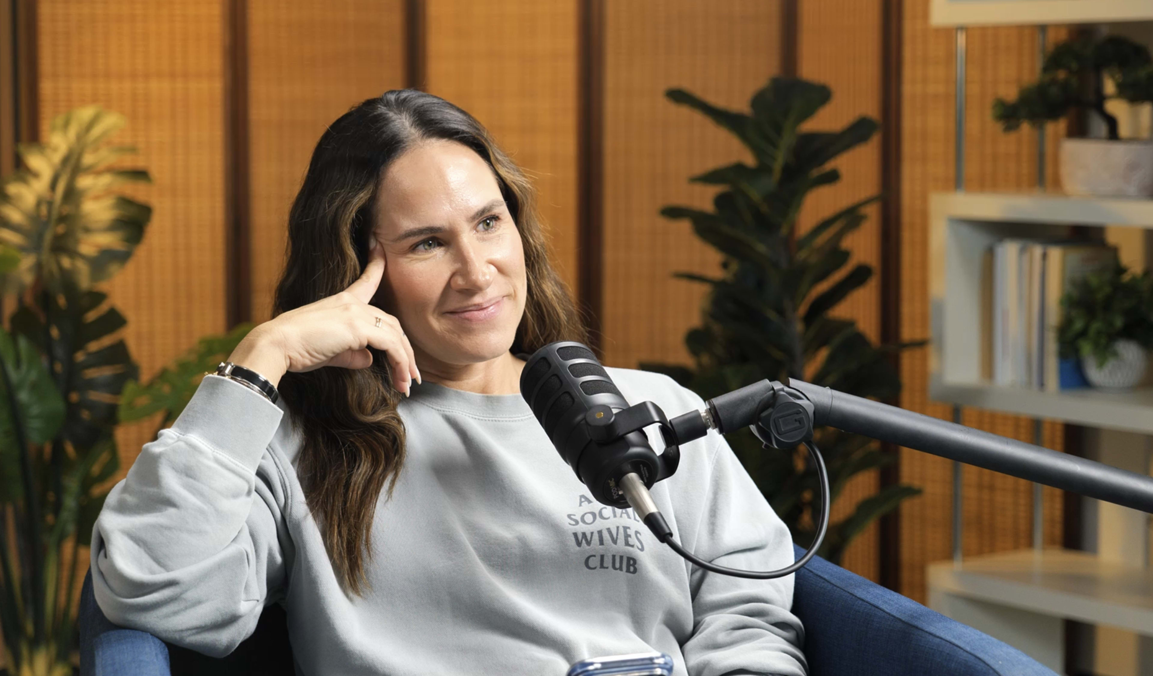 A woman sits thoughtfully in a chair, wearing a sweatshirt and looking at a microphone in front of her during a podcast recording.