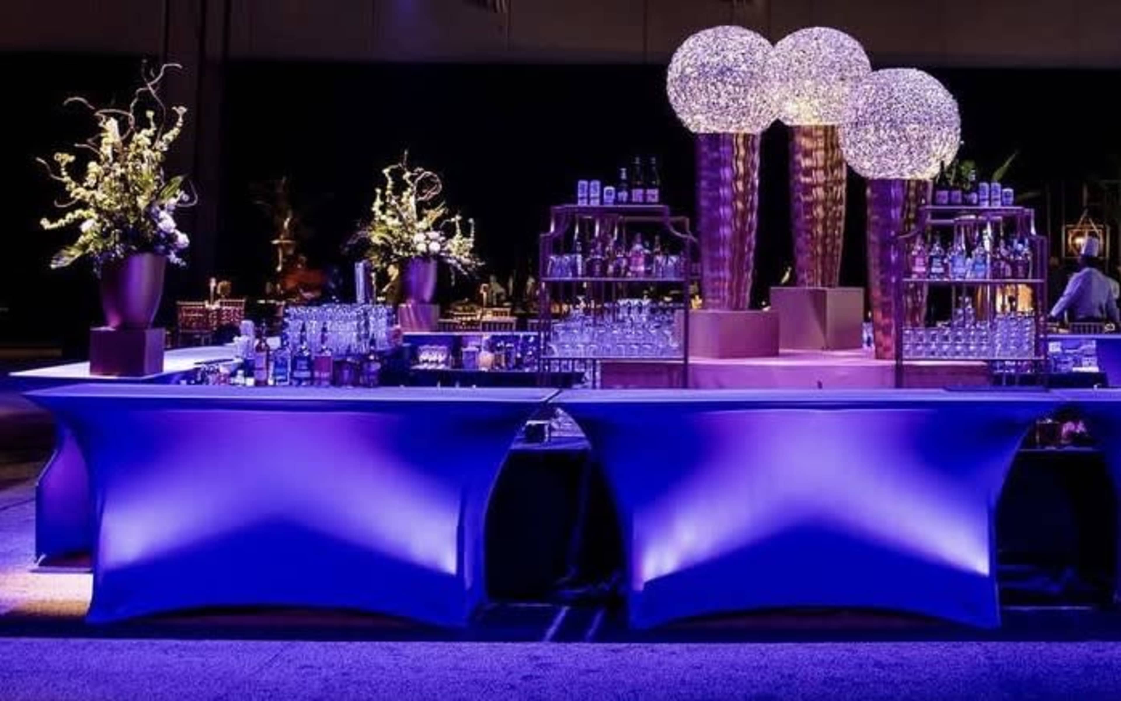 The image shows a elegantly designed bar setup with illuminated tables, floral arrangements, and ornate light fixtures.