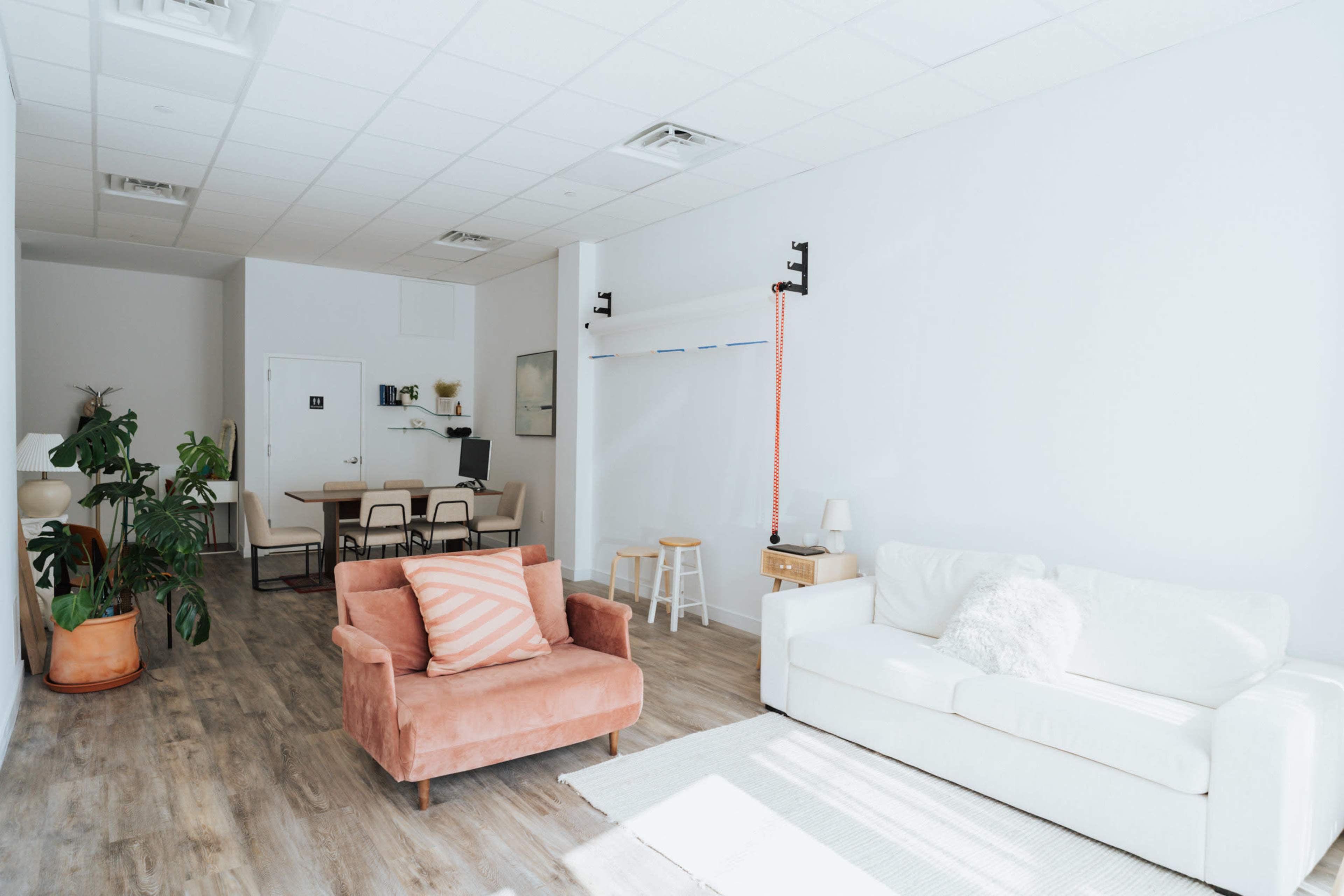 A bright, minimalist interior with a pink armchair, a white sofa, a dining area in the background, and large windows letting in natural light.