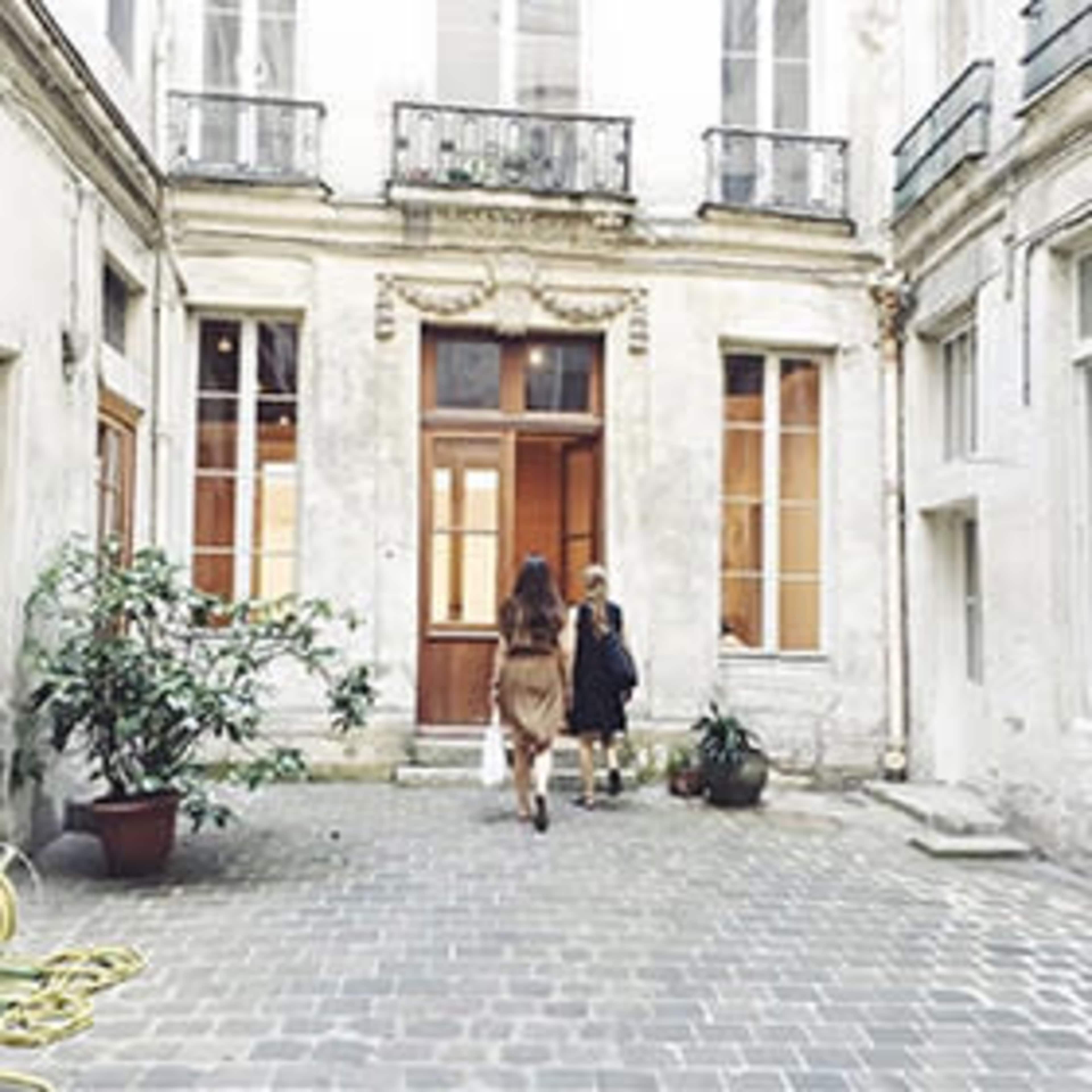 Two women walk toward a wooden door in a courtyard with stone paving and potted plants.