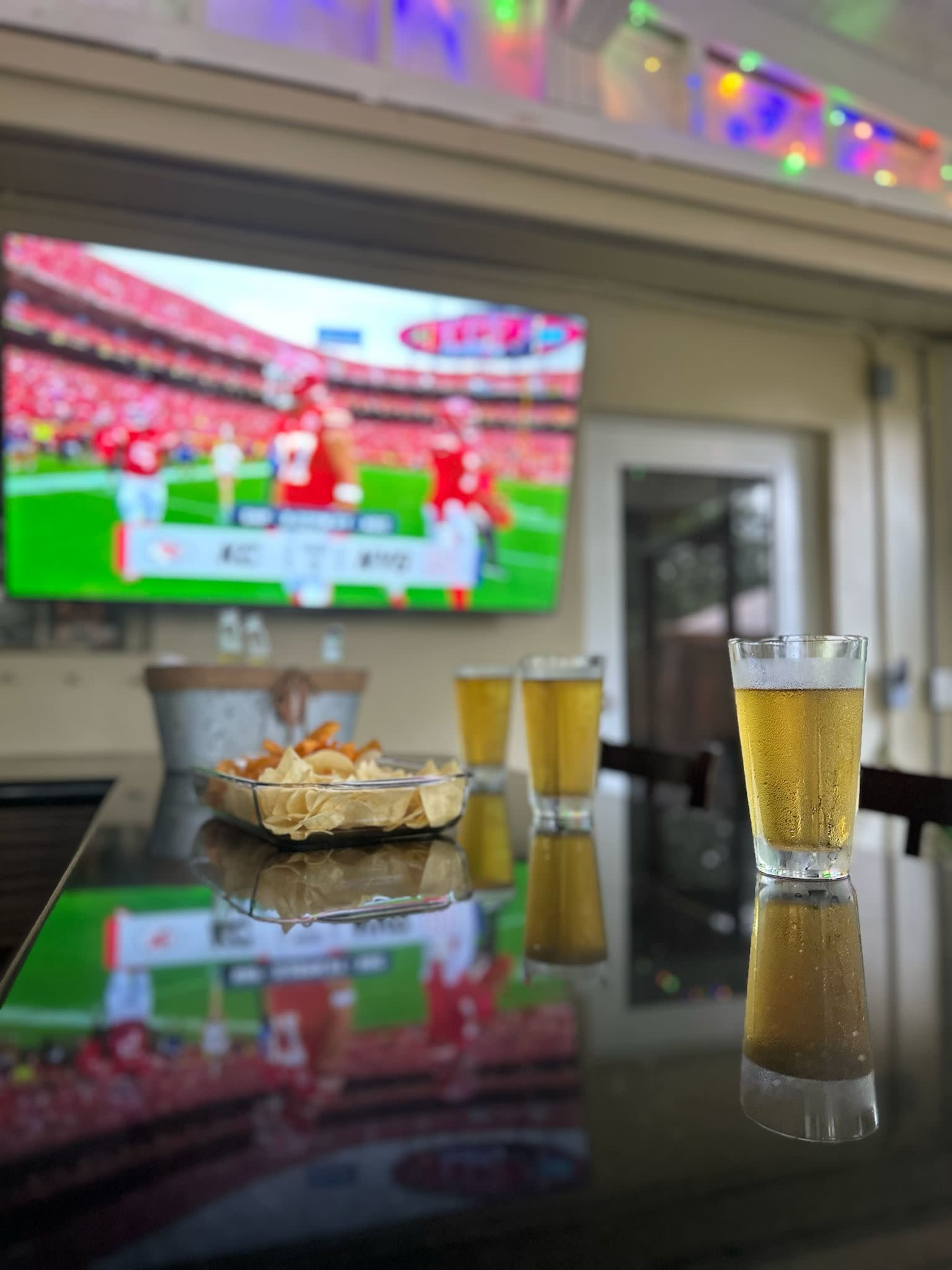 The image shows a bar counter with glasses of beer and a plate of snacks in front of a television displaying a sports game.