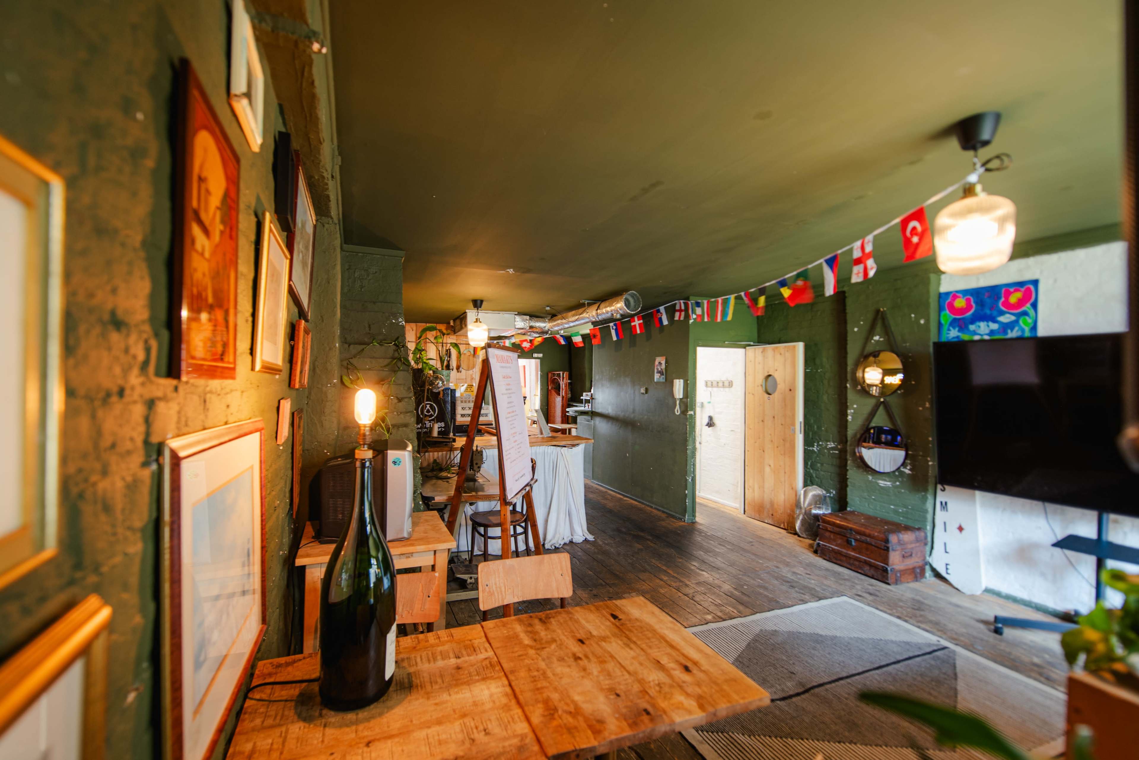 The image shows a cozy interior space with green walls, wooden furniture, and various framed artworks, adorned with flags hanging from the ceiling.