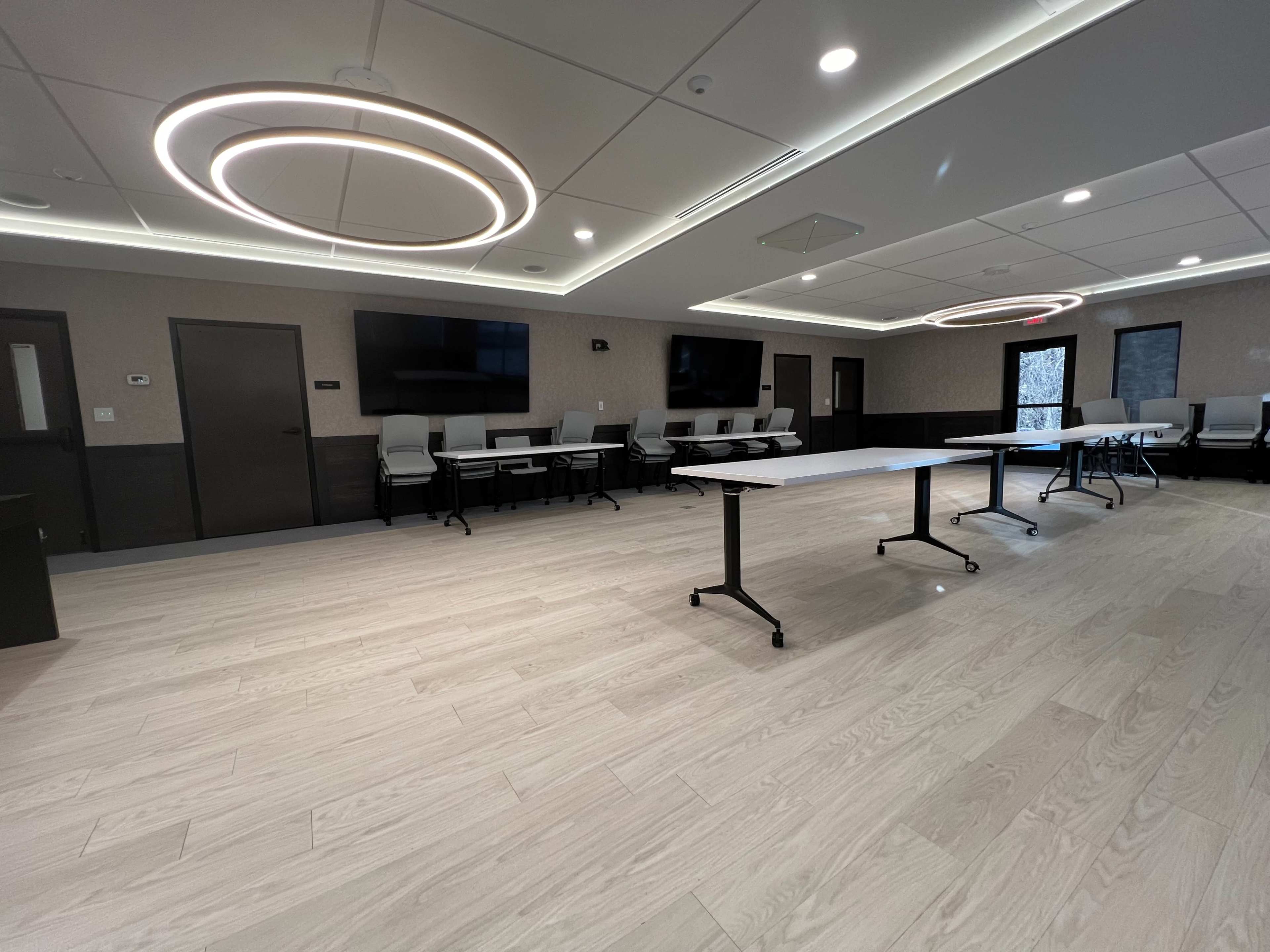 A modern meeting room with light wood flooring, two circular overhead lights, and tables arranged in the center.