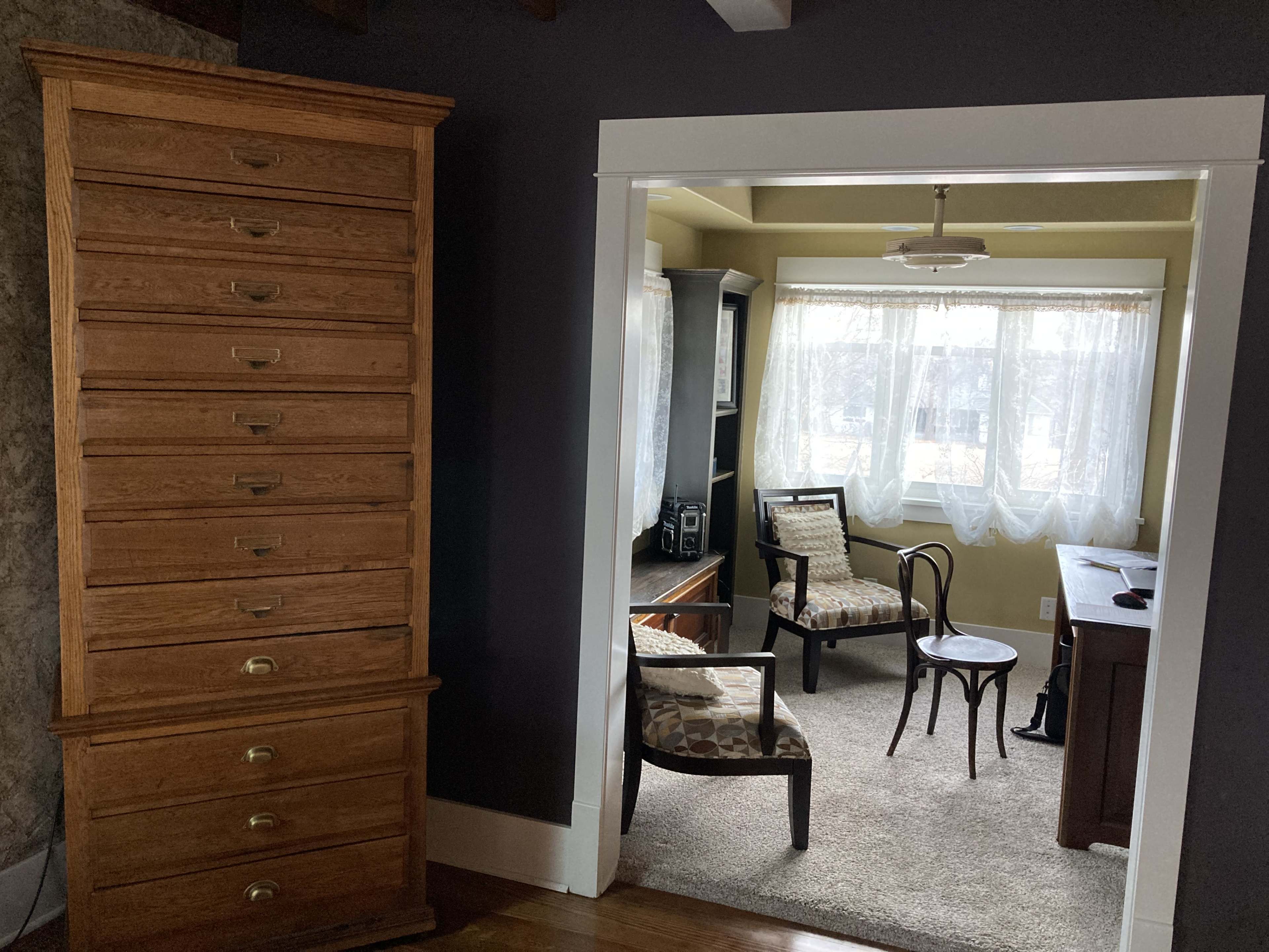 A tall, wooden chest of drawers stands beside an open doorway leading to a small, brightly lit sitting area.
