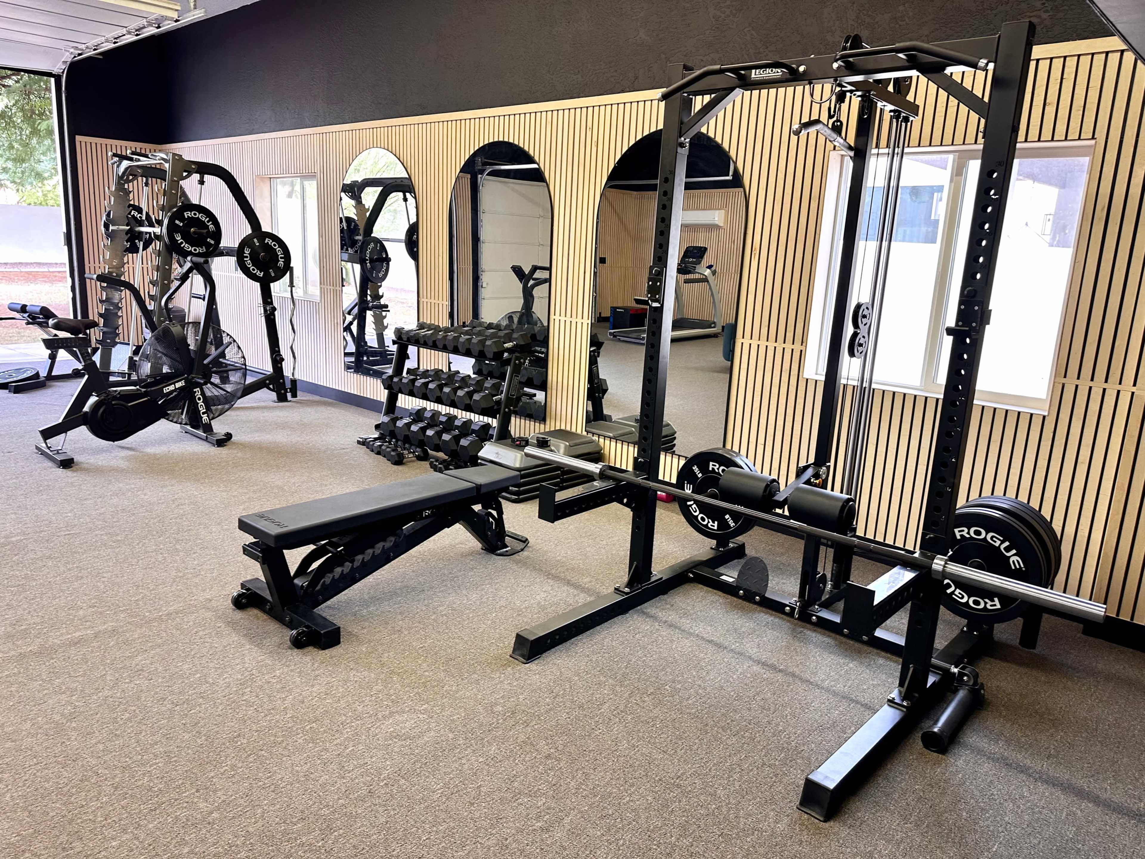 The image shows a gym interior featuring weightlifting equipment, including a bench press, cable machine, and free weights, with mirrors on the wall and cardio machines in the background.