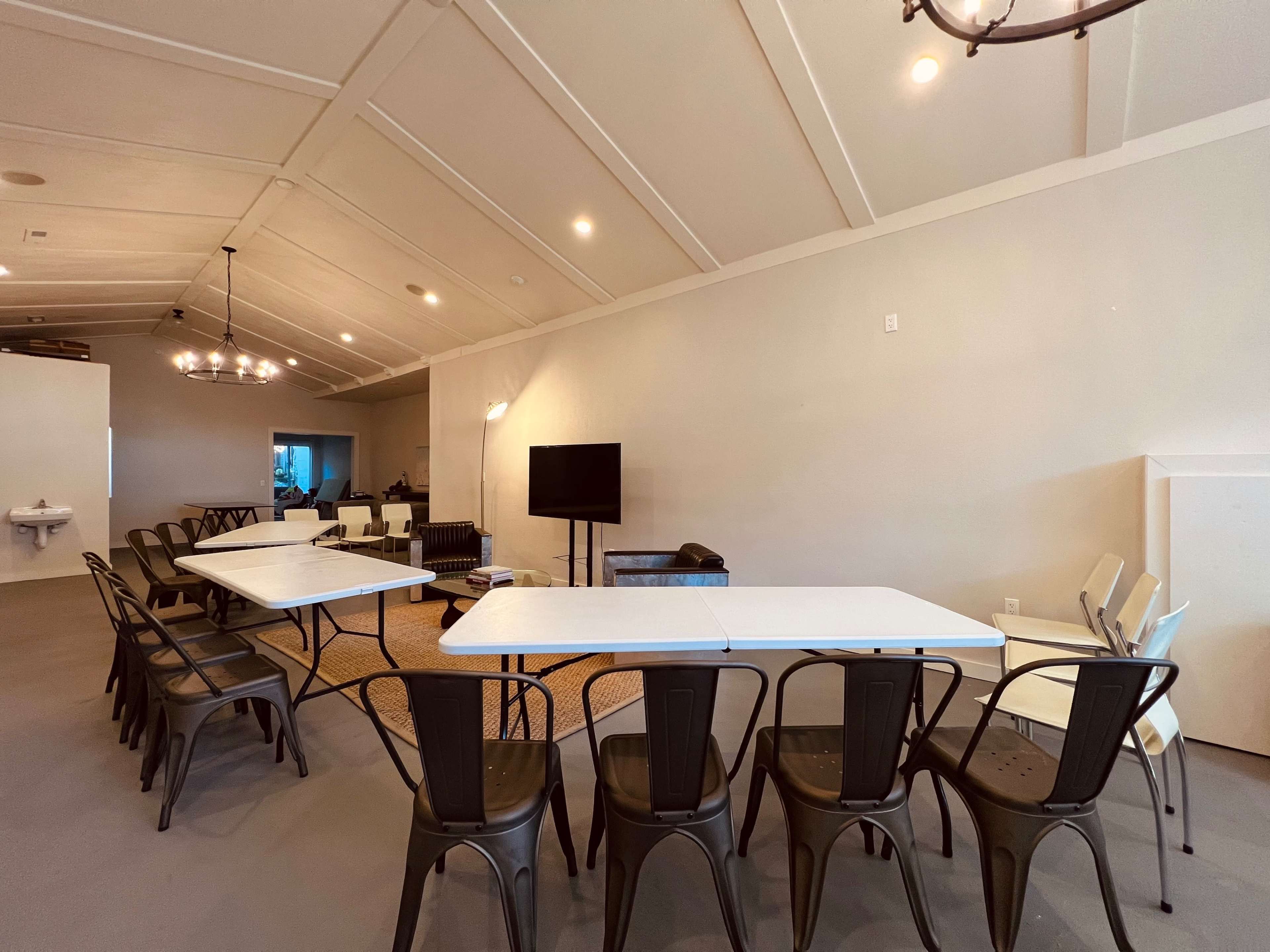 A large meeting room with multiple tables arranged in a U-shape and chairs placed around them, featuring a television and a seating area in the background.