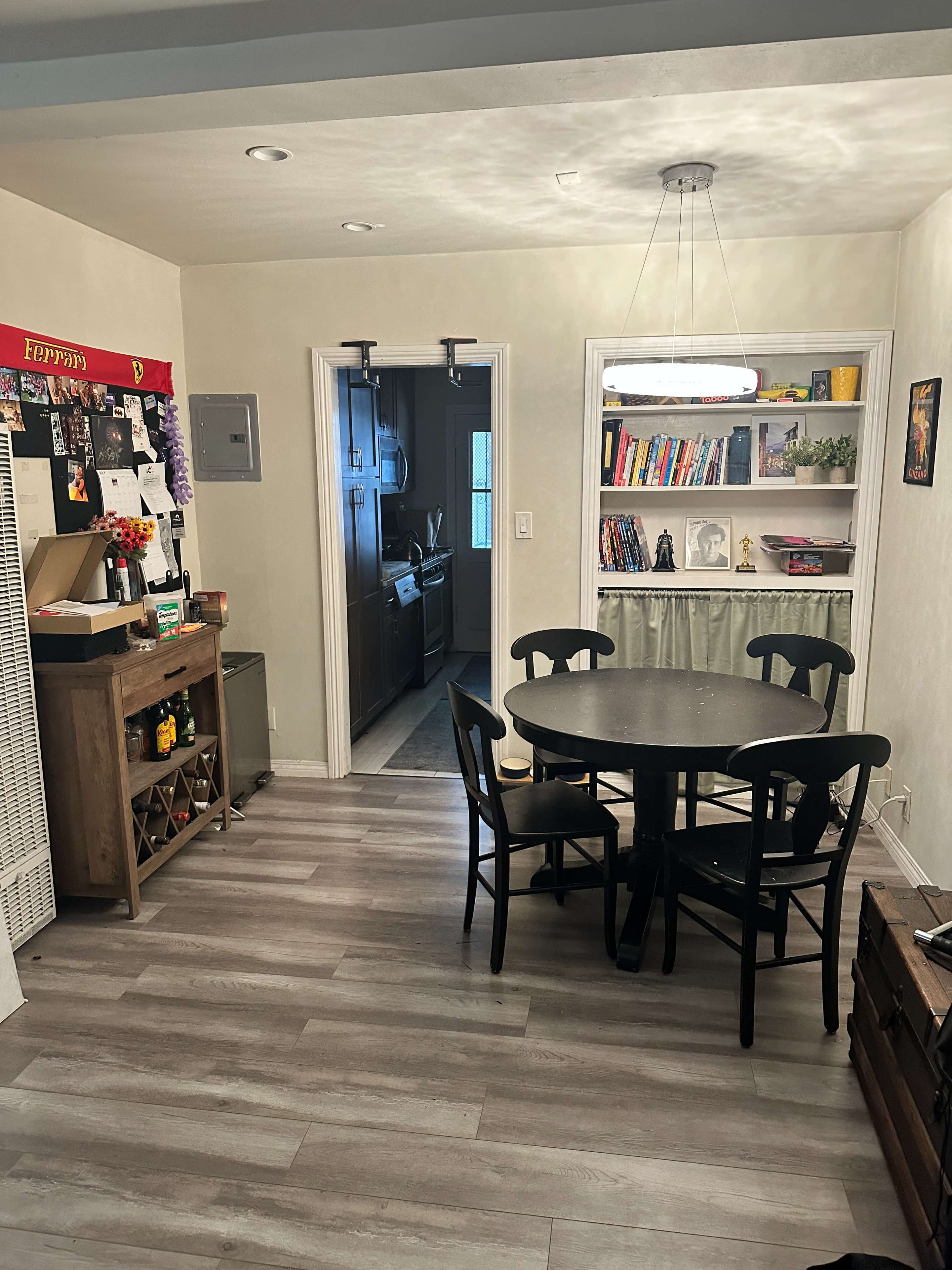 The image shows a well-lit kitchen area featuring a round black table with chairs, a wooden sideboard, and a doorway leading to a darker kitchen.