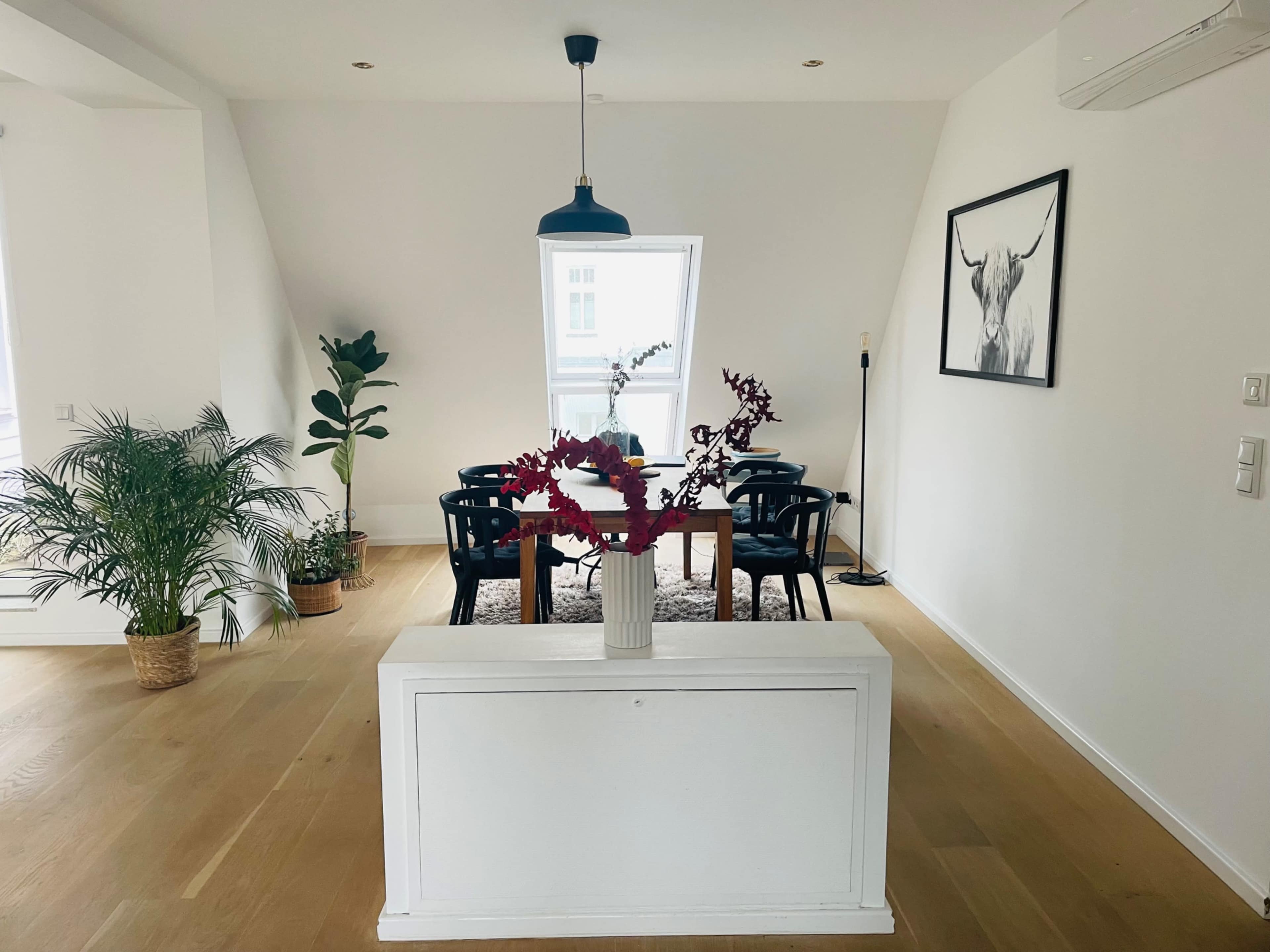 A bright, minimalist living space with a dining area, a large window, plants, and a cow artwork on the wall.