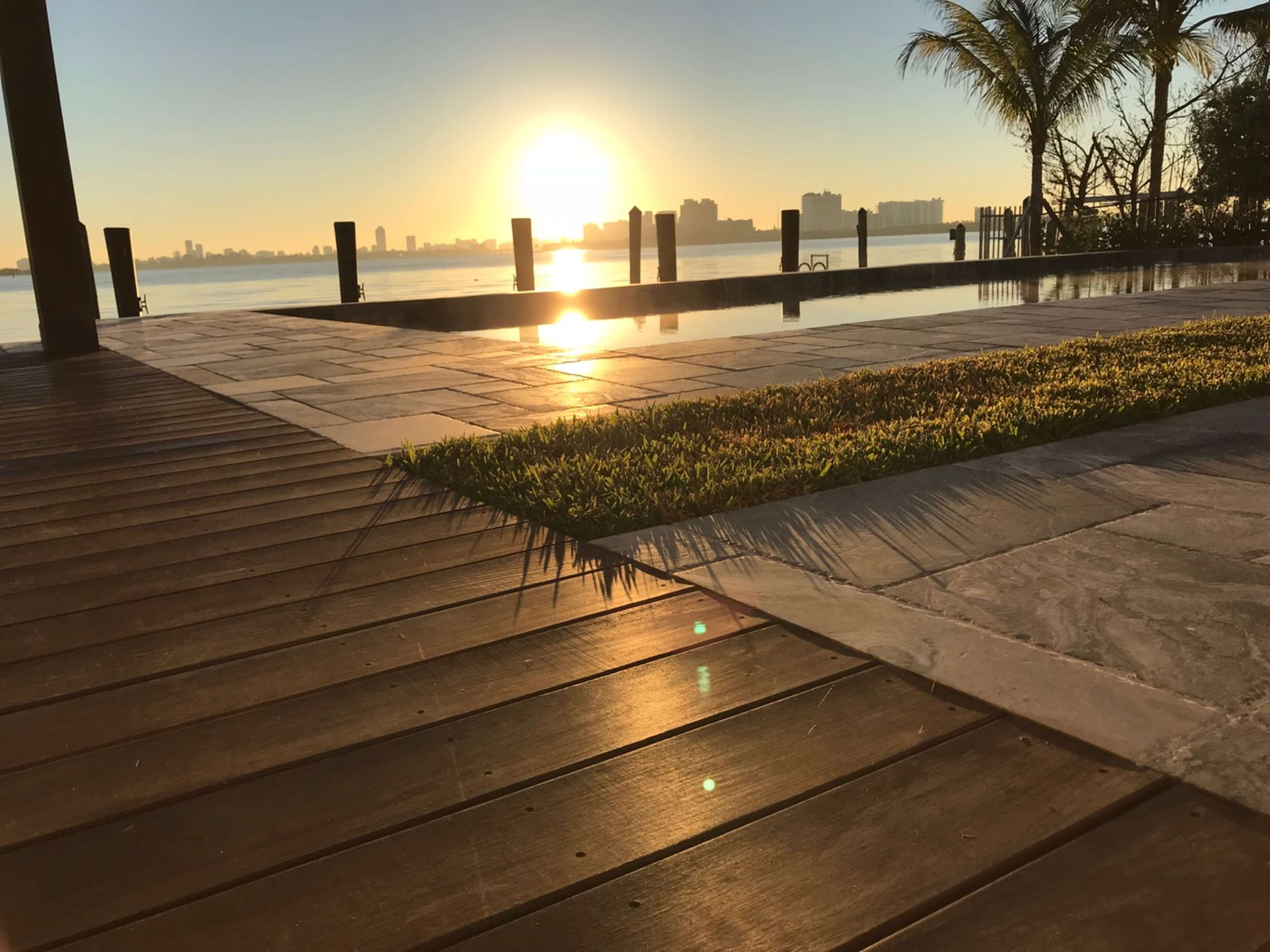 The image shows a wooden deck leading to a pool area with a view of a sunrise over a calm water surface and city skyline in the background.