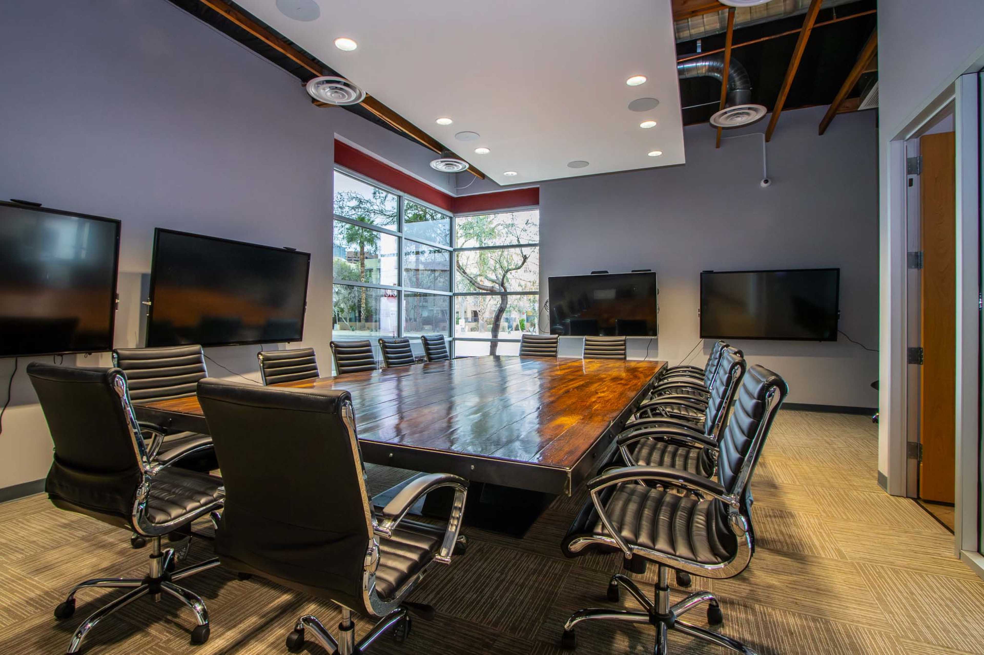 A modern conference room features a large wooden table surrounded by black leather chairs, with multiple screens mounted on the walls.