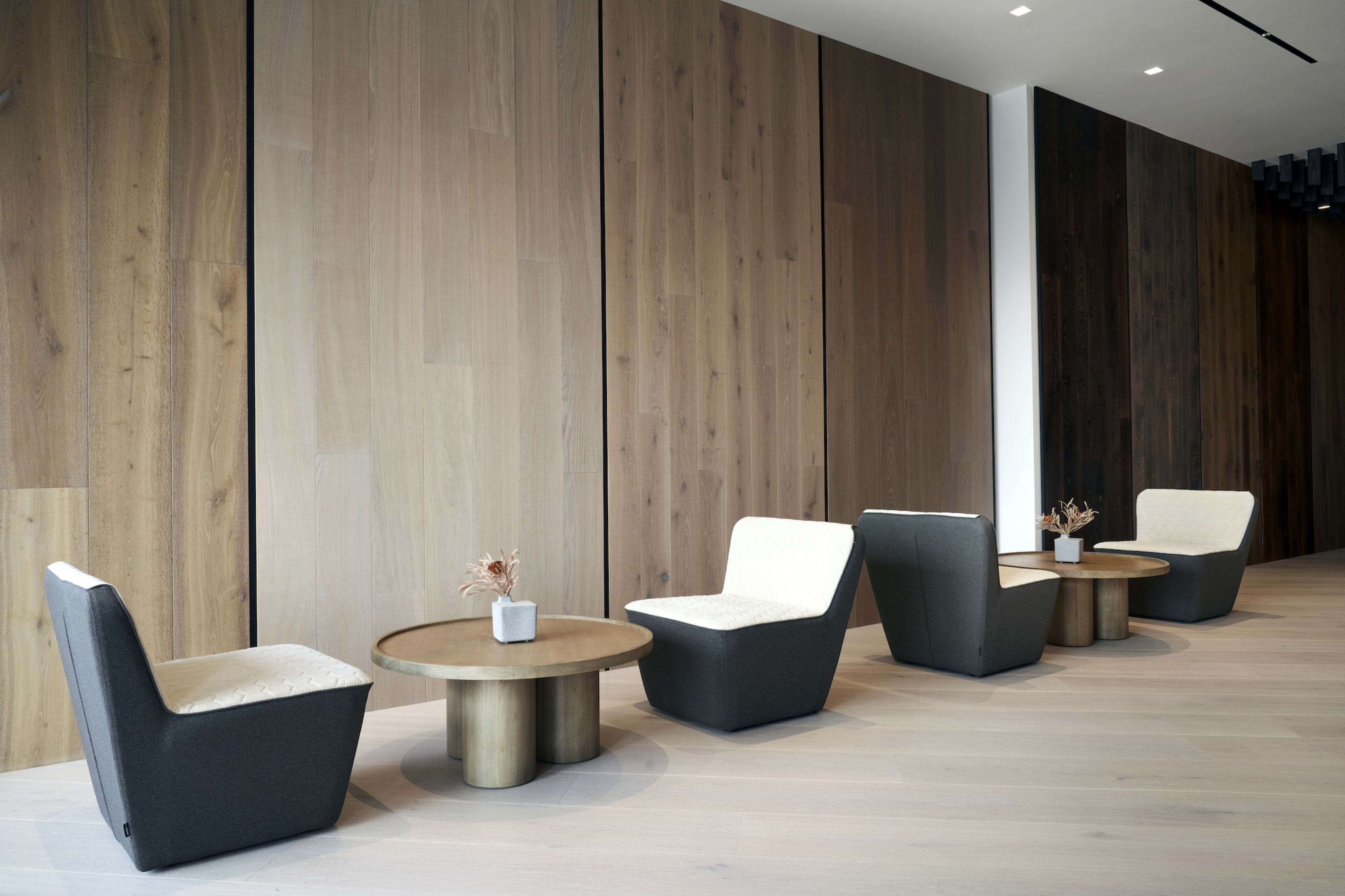 A modern waiting area featuring four upholstered chairs arranged around two round wooden tables, with a wooden accent wall in the background.