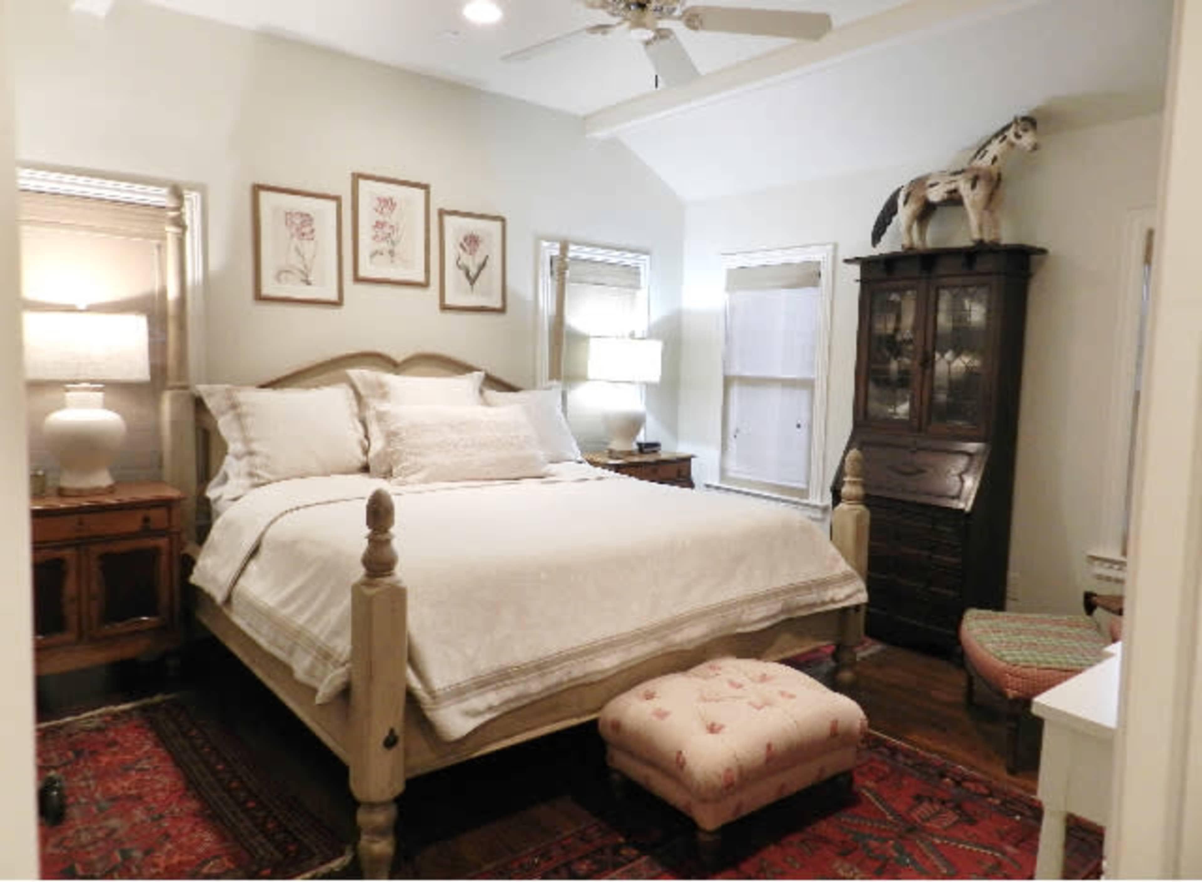 The image shows a cozy bedroom featuring a large bed with white bedding, two bedside lamps, a wooden dresser, and decorative wall art.