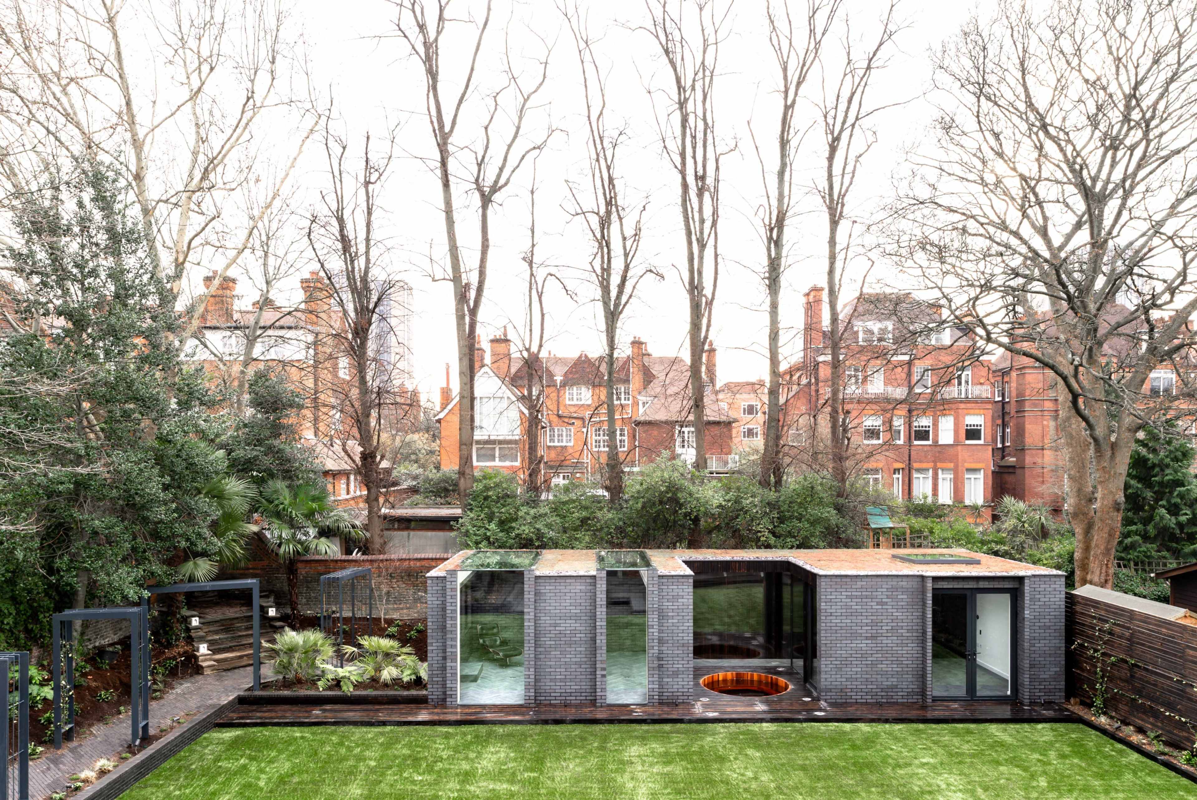 The image shows a modern, low-rise building with large windows and a green lawn, surrounded by bare trees and traditional residential buildings in the background.