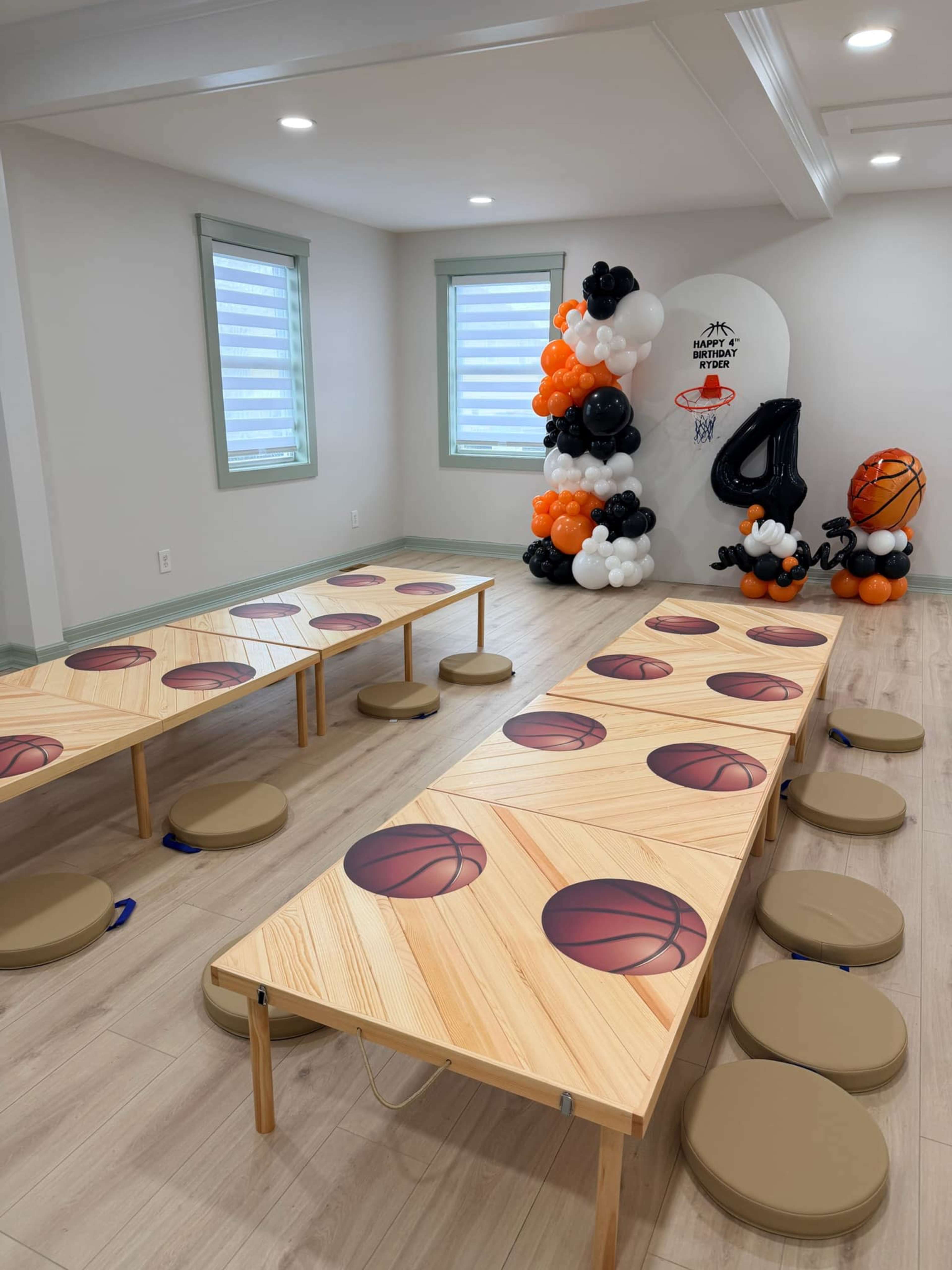 The image shows a decorated room with two tables featuring basketball-themed designs and circular stools arranged around them, along with balloon decorations in the corner.