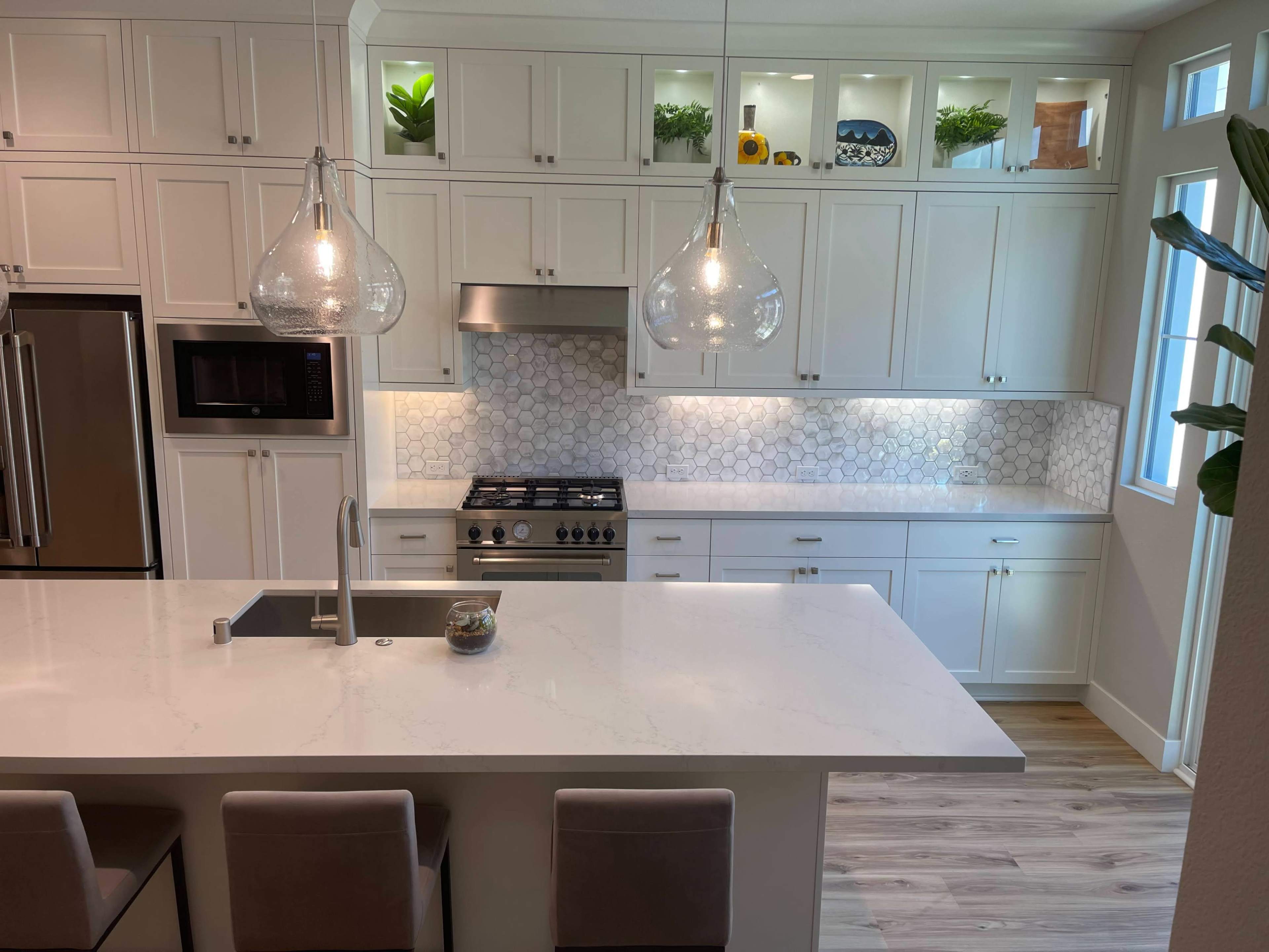 A modern kitchen featuring white cabinetry, a large island with a sleek countertop, and pendant lights above, accompanied by a tiled backsplash and stainless steel appliances.