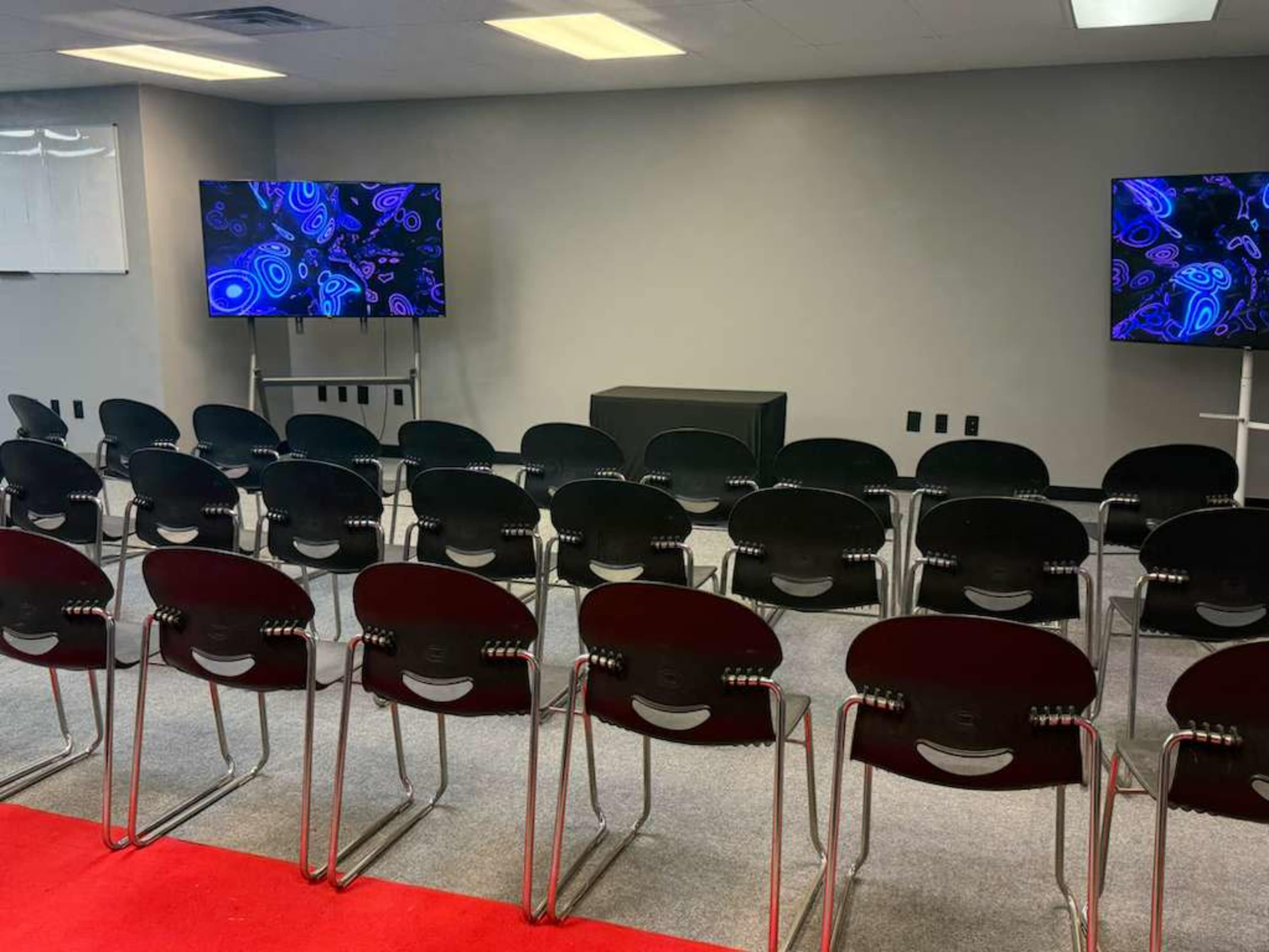 The image shows a conference room with several rows of black chairs facing two screens displaying abstract colorful graphics.