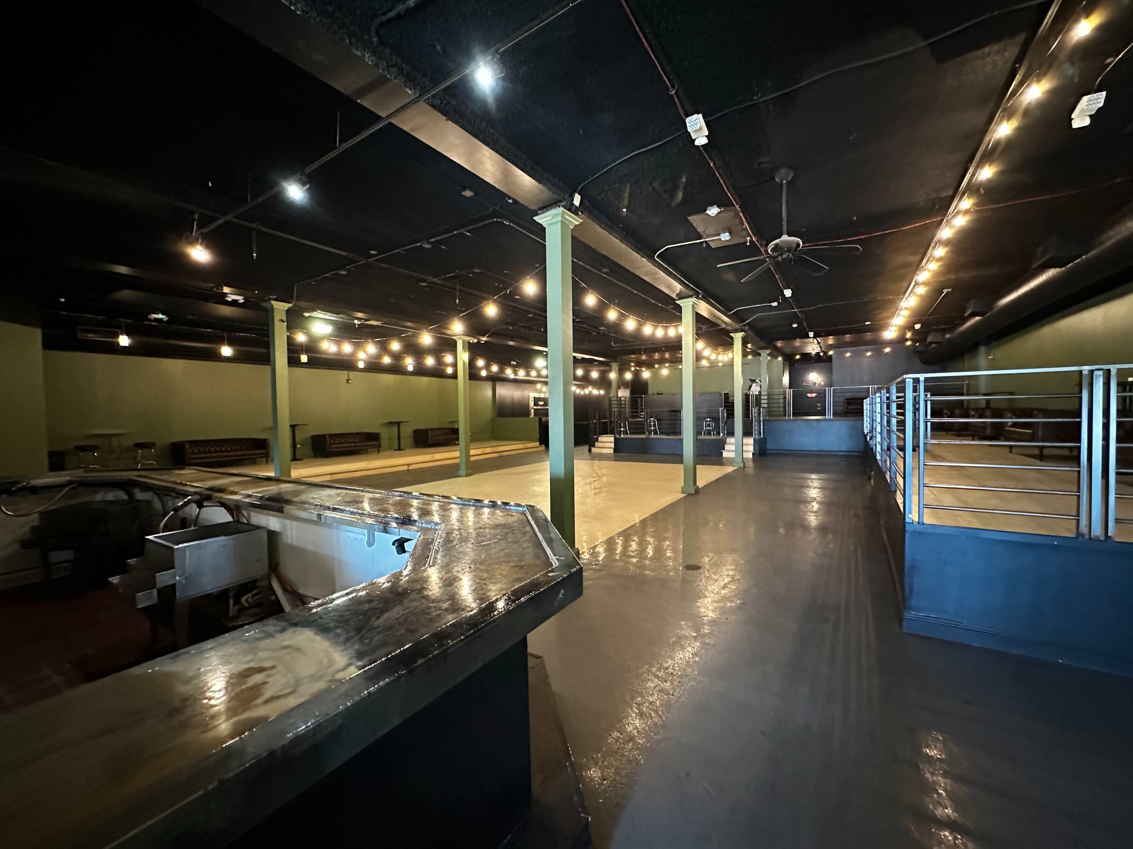 The image shows an empty interior of a venue with a polished floor, exposed lighting, and a bar area in the foreground.