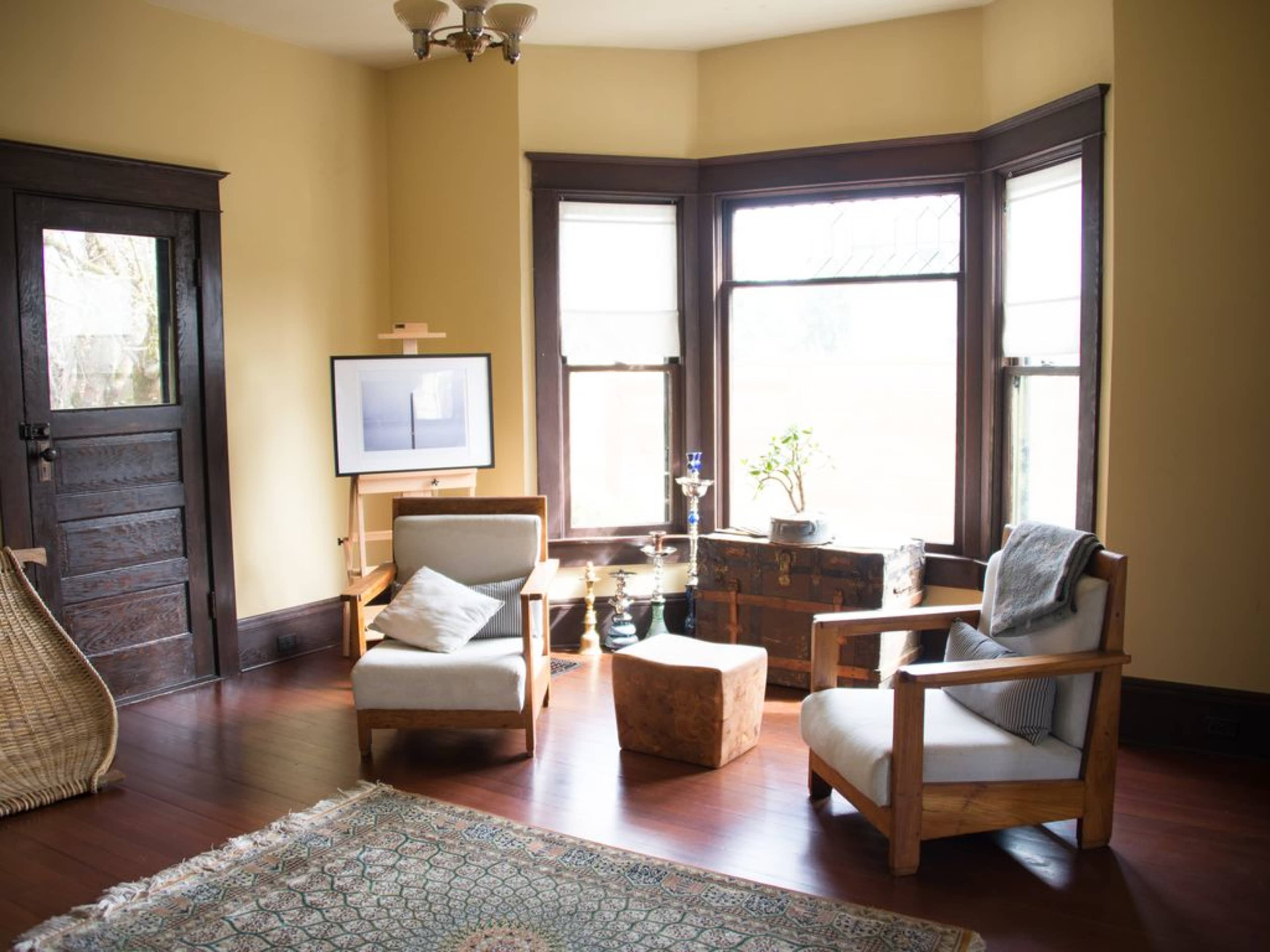 A cozy living room with two wooden chairs, a small ottoman, a flat-screen TV on an easel, and large windows allowing natural light.