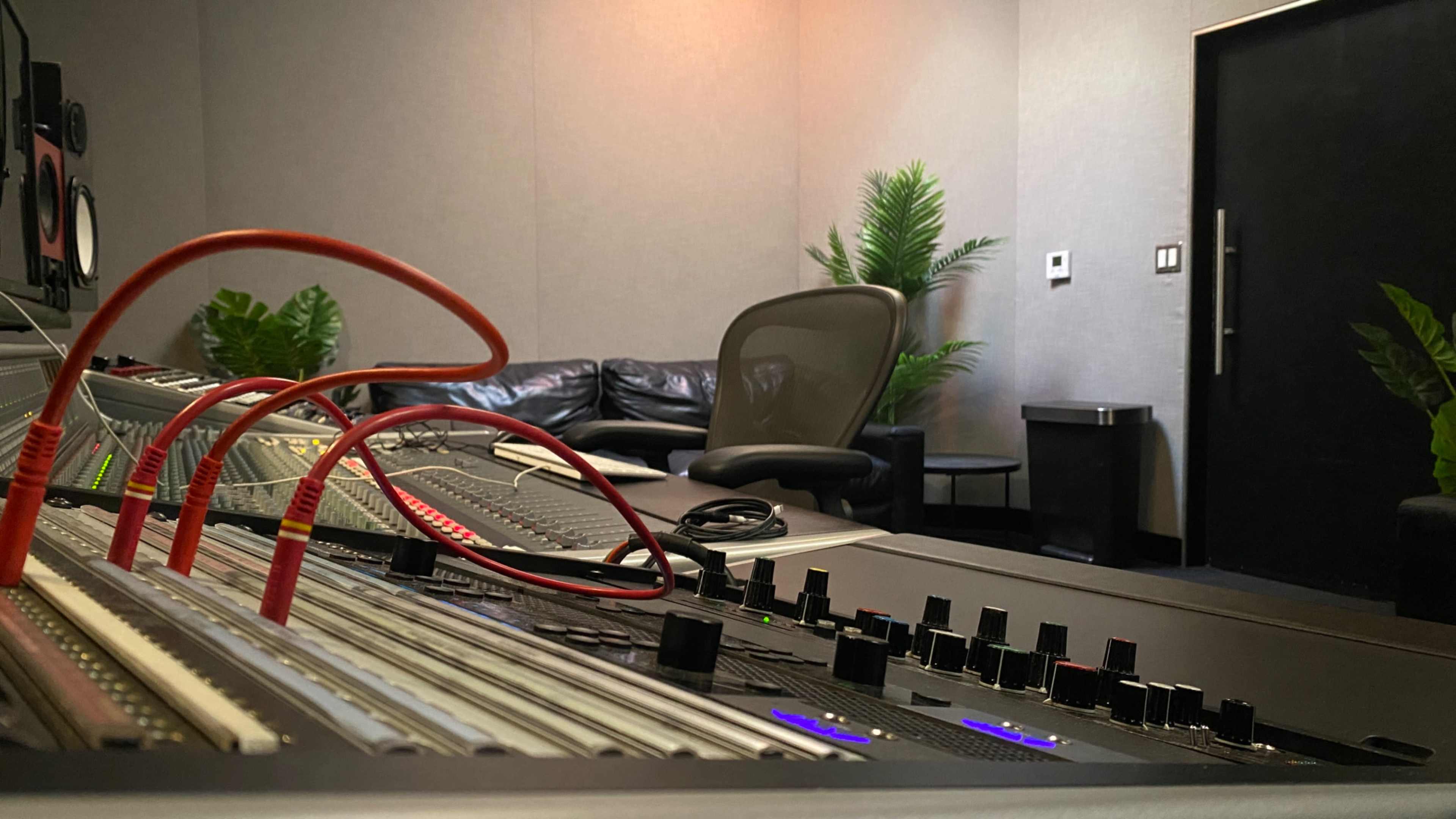 The image shows a sound mixing console with cables in the foreground, and a couch and potted plants in the background of a recording studio.