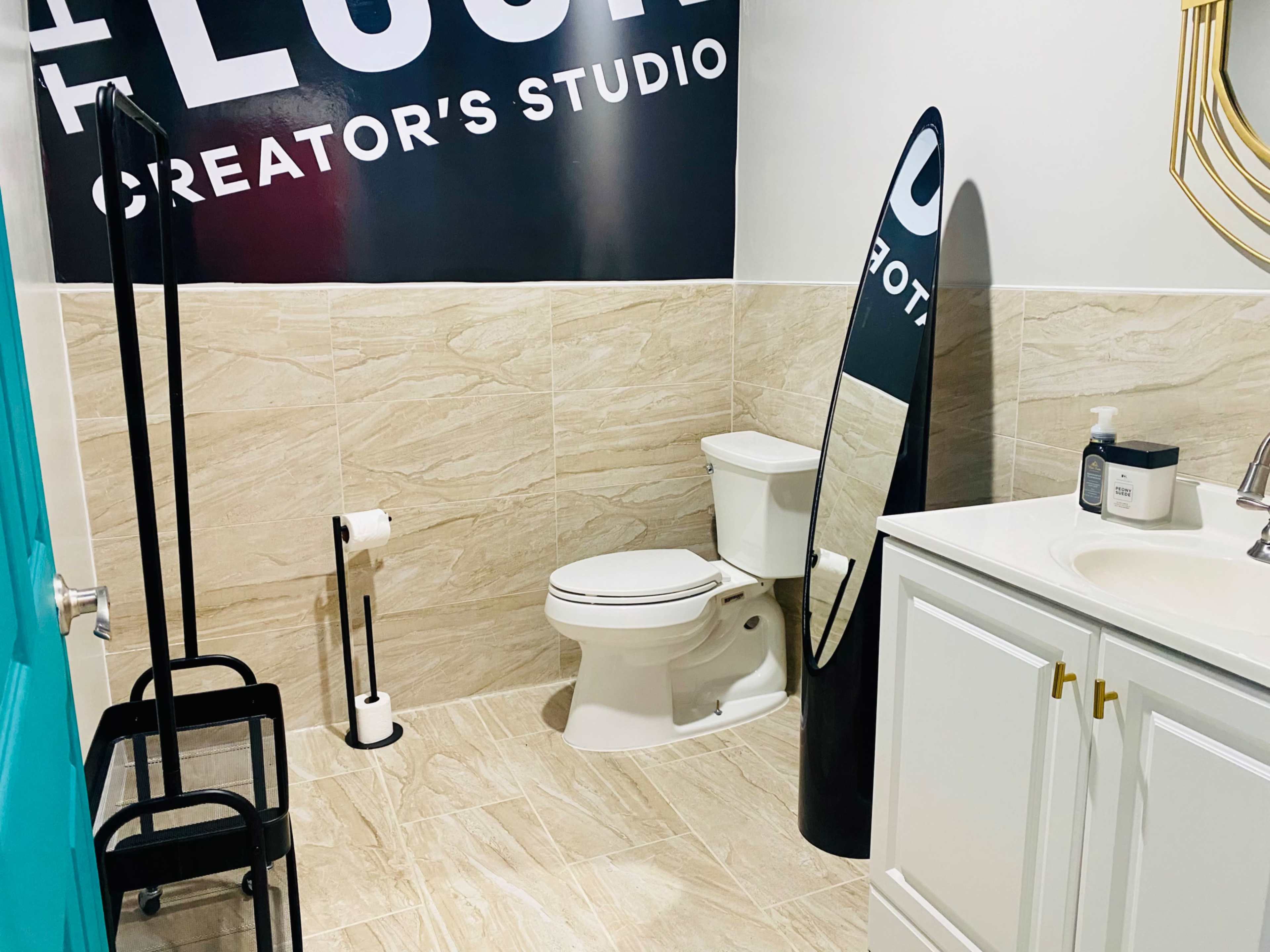 The image shows a bathroom featuring a toilet, a mirror, a small sink with storage cabinets, and a wall with the text "Creator's Studio."