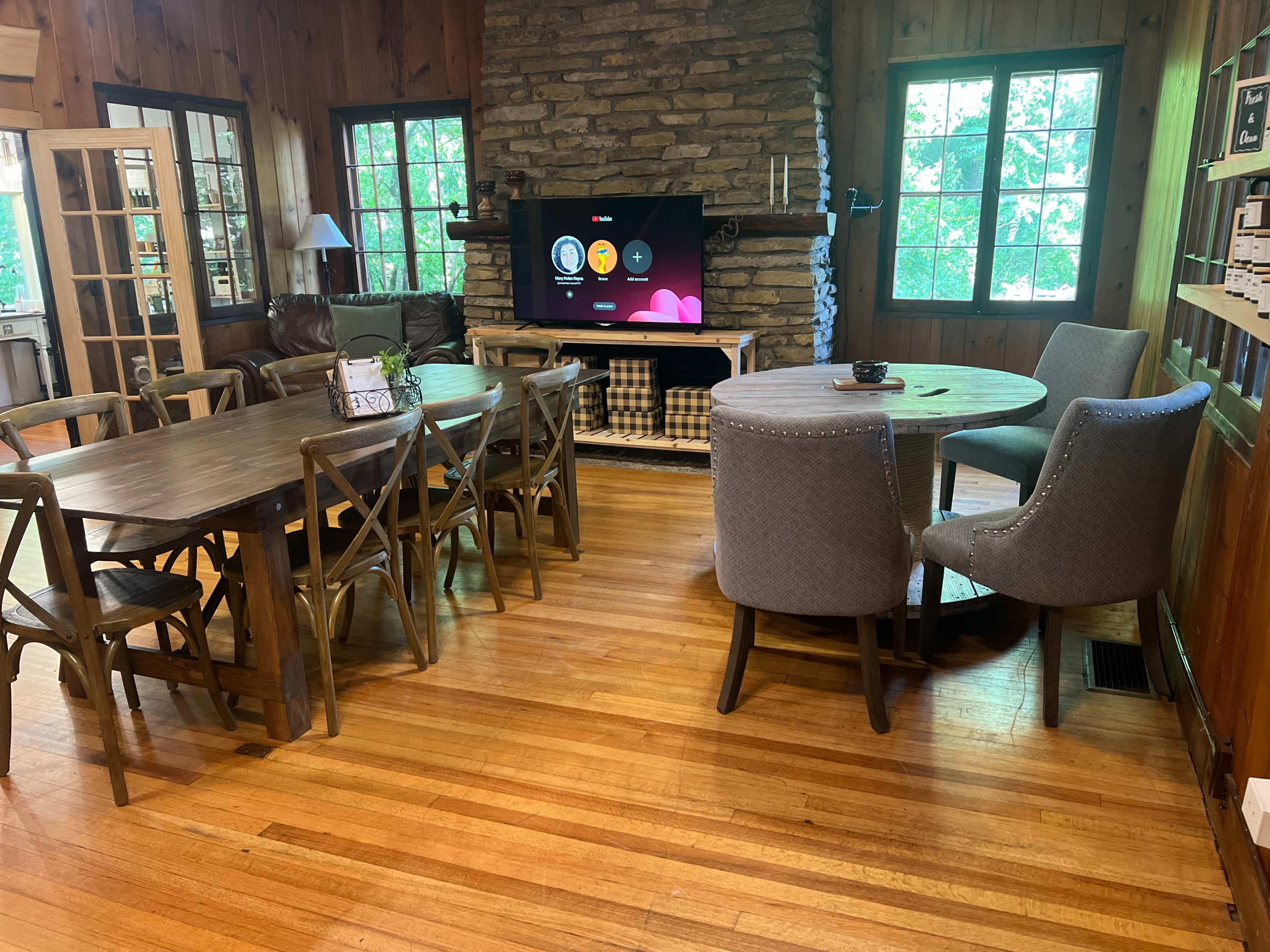 The image shows a cozy wooden interior featuring a dining area with a large table and chairs, a living space with a television mounted on a stone wall, and large windows letting in natural light.