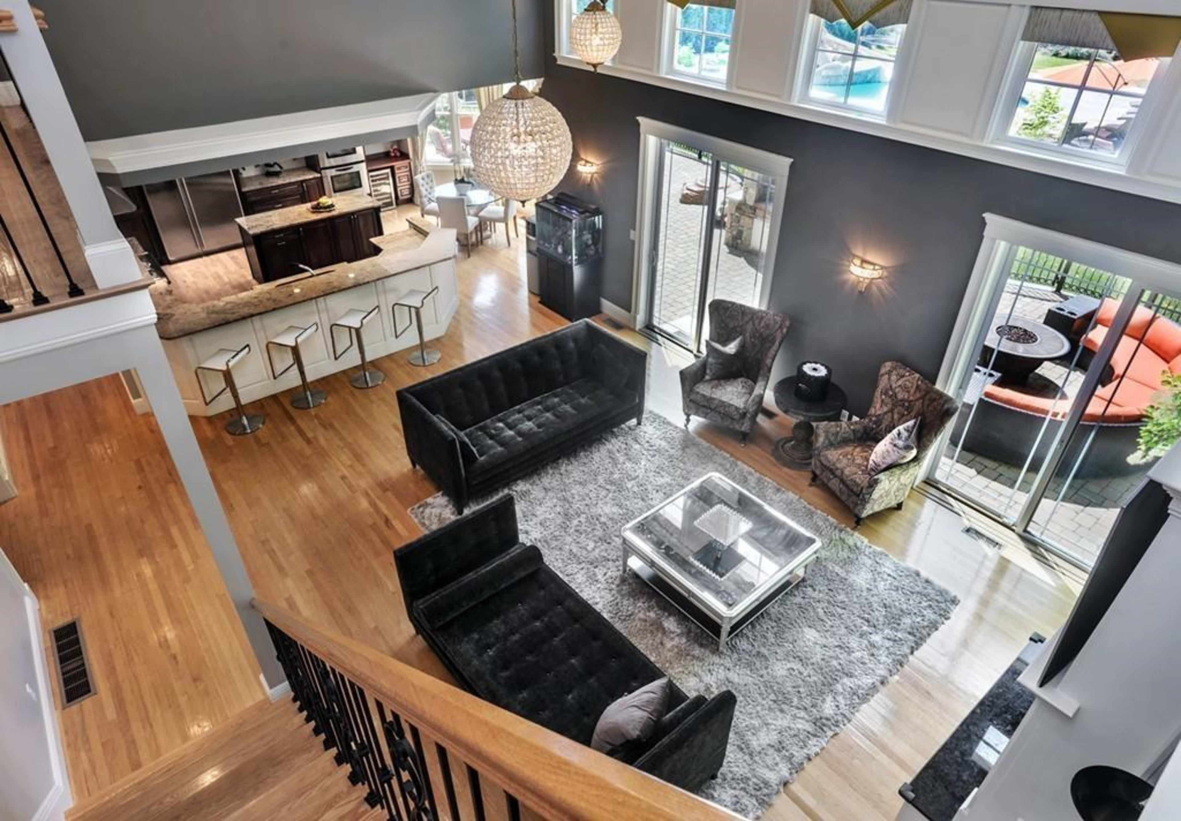 The image shows a modern open-concept living room with a view of a kitchen, featuring two dark sofas, two armchairs, a glass coffee table, and large windows overlooking a patio.
