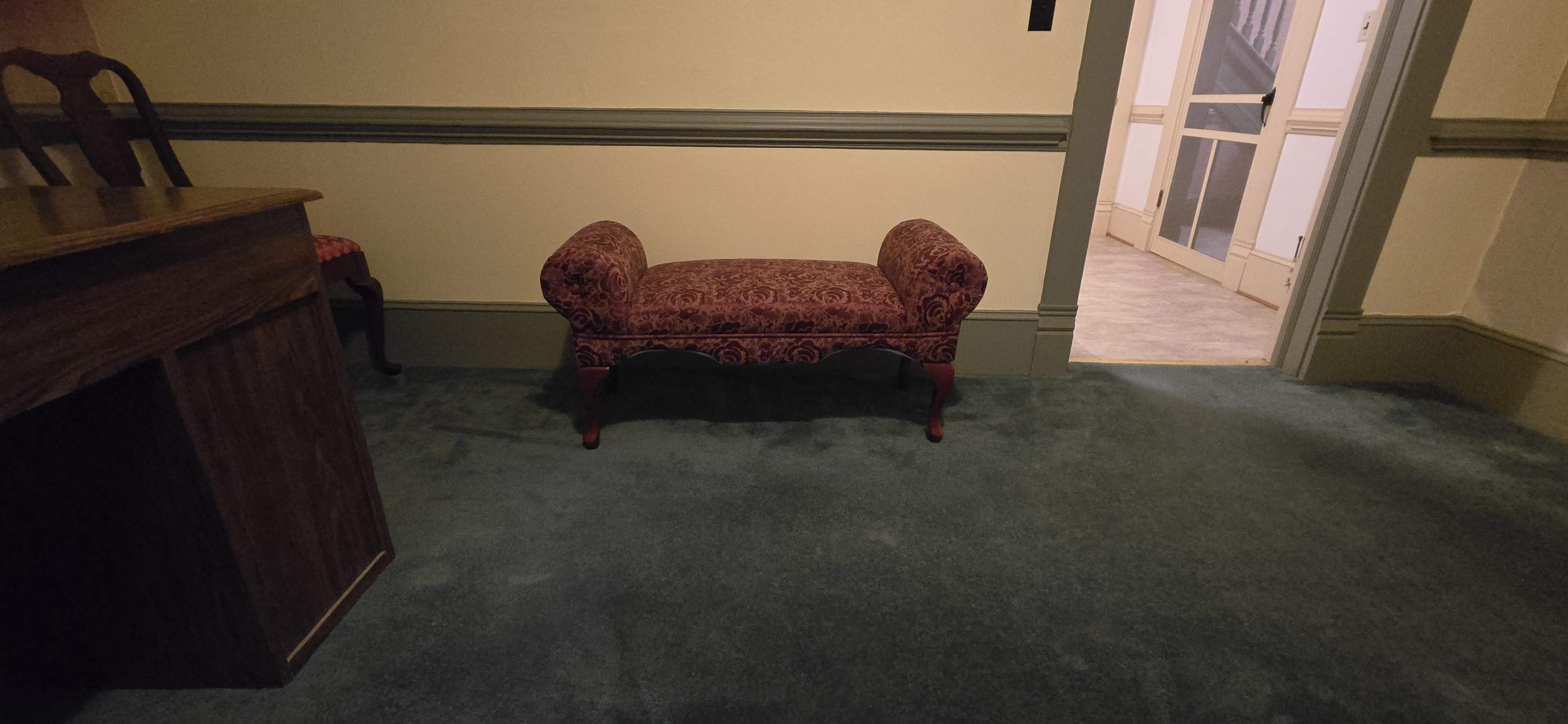 The image shows a room with green carpet, a patterned red bench in the center, and a doorway leading to another area.