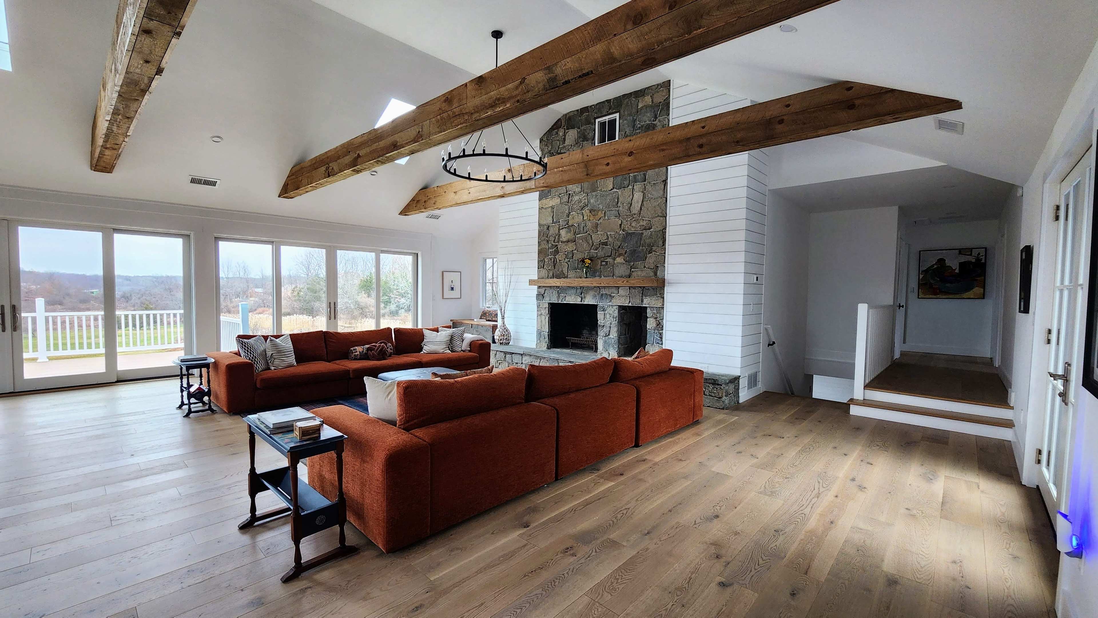 The living room features a large orange sectional sofa facing a stone fireplace beneath exposed wooden beams, with sliding glass doors leading to a view outside.