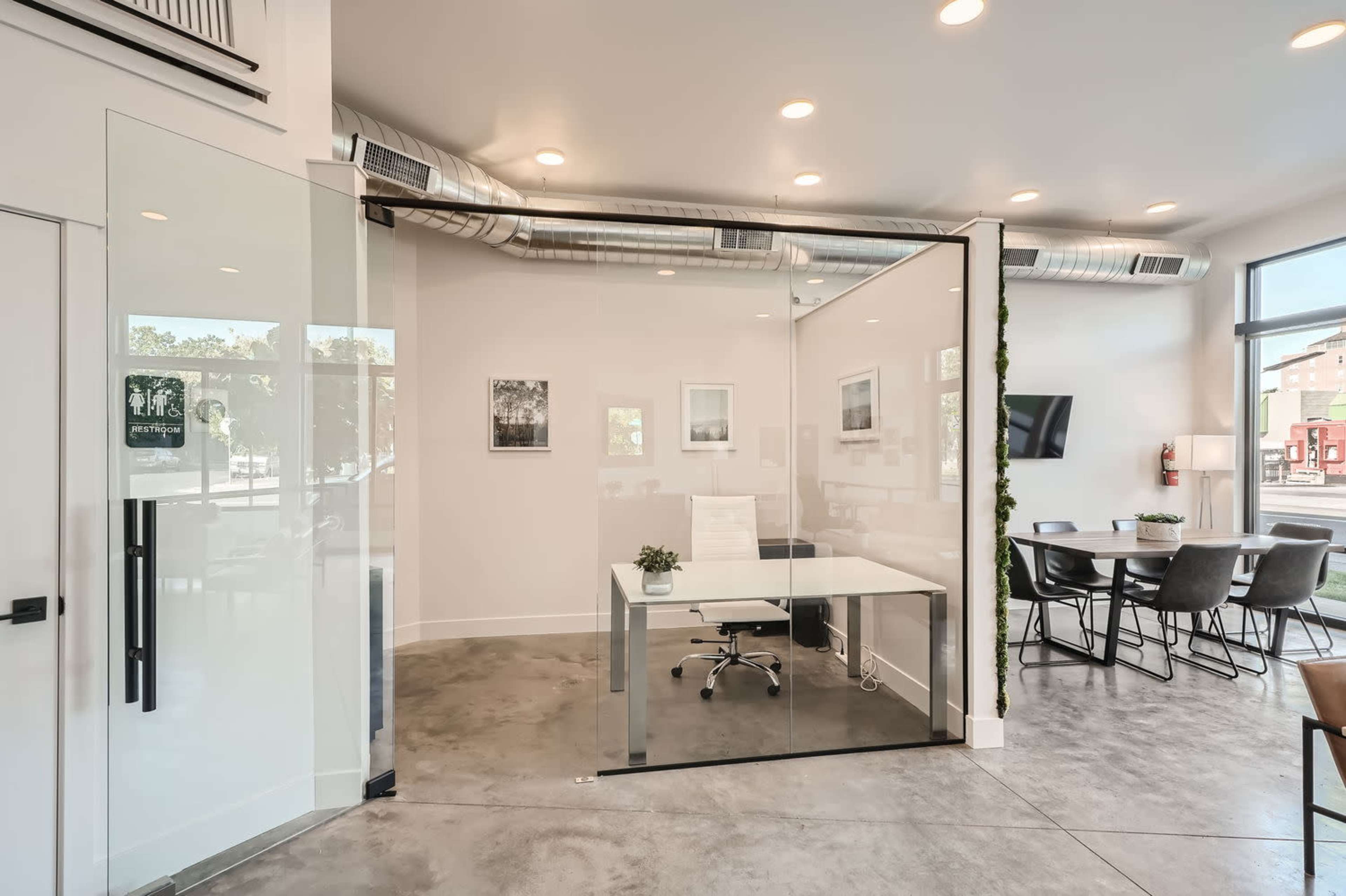 The image shows a modern office space with a glass-enclosed meeting room, a desk with a chair, and a seating area, all featuring industrial-style decor.