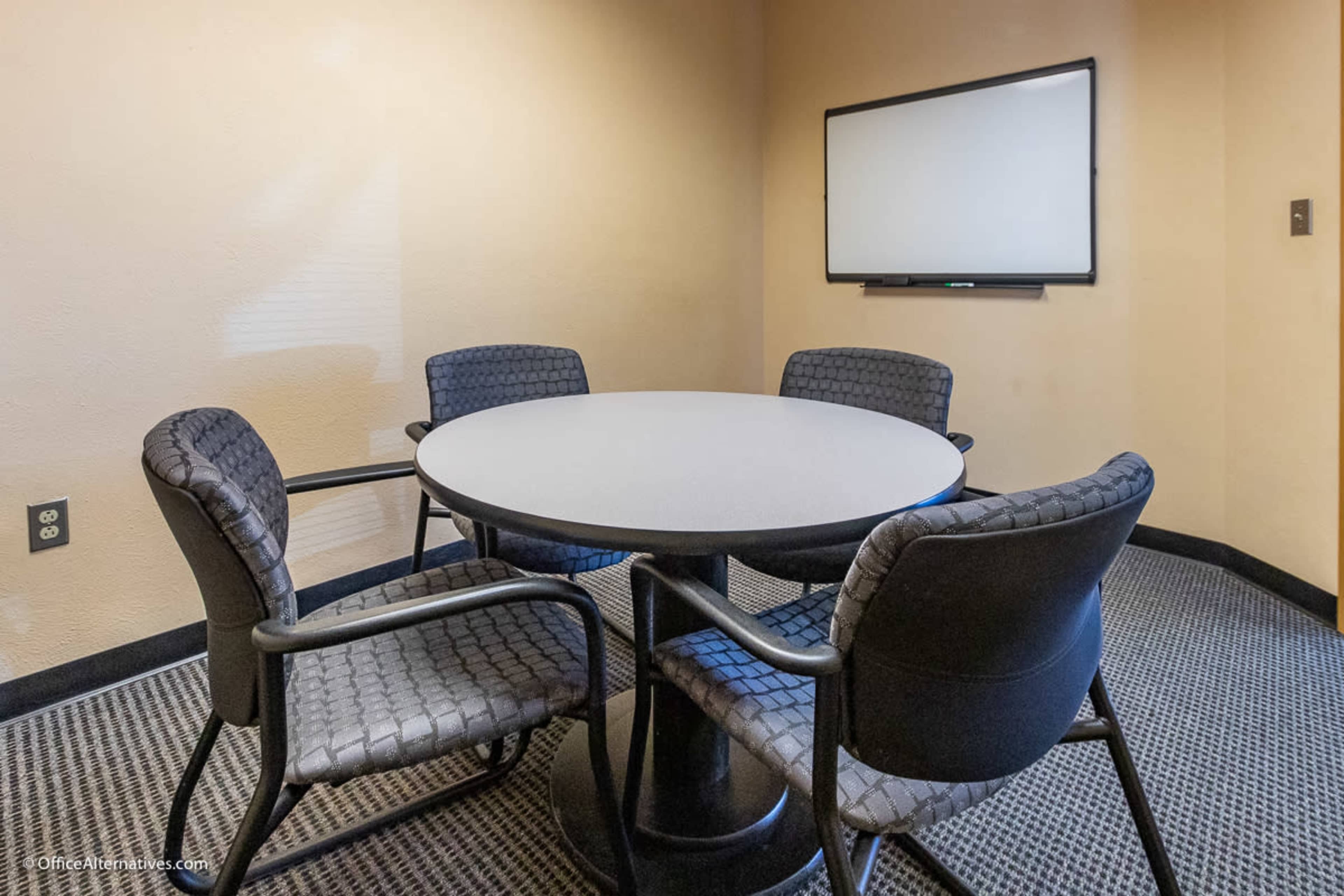 A small conference room features a round table with four black chairs and a wall-mounted whiteboard.