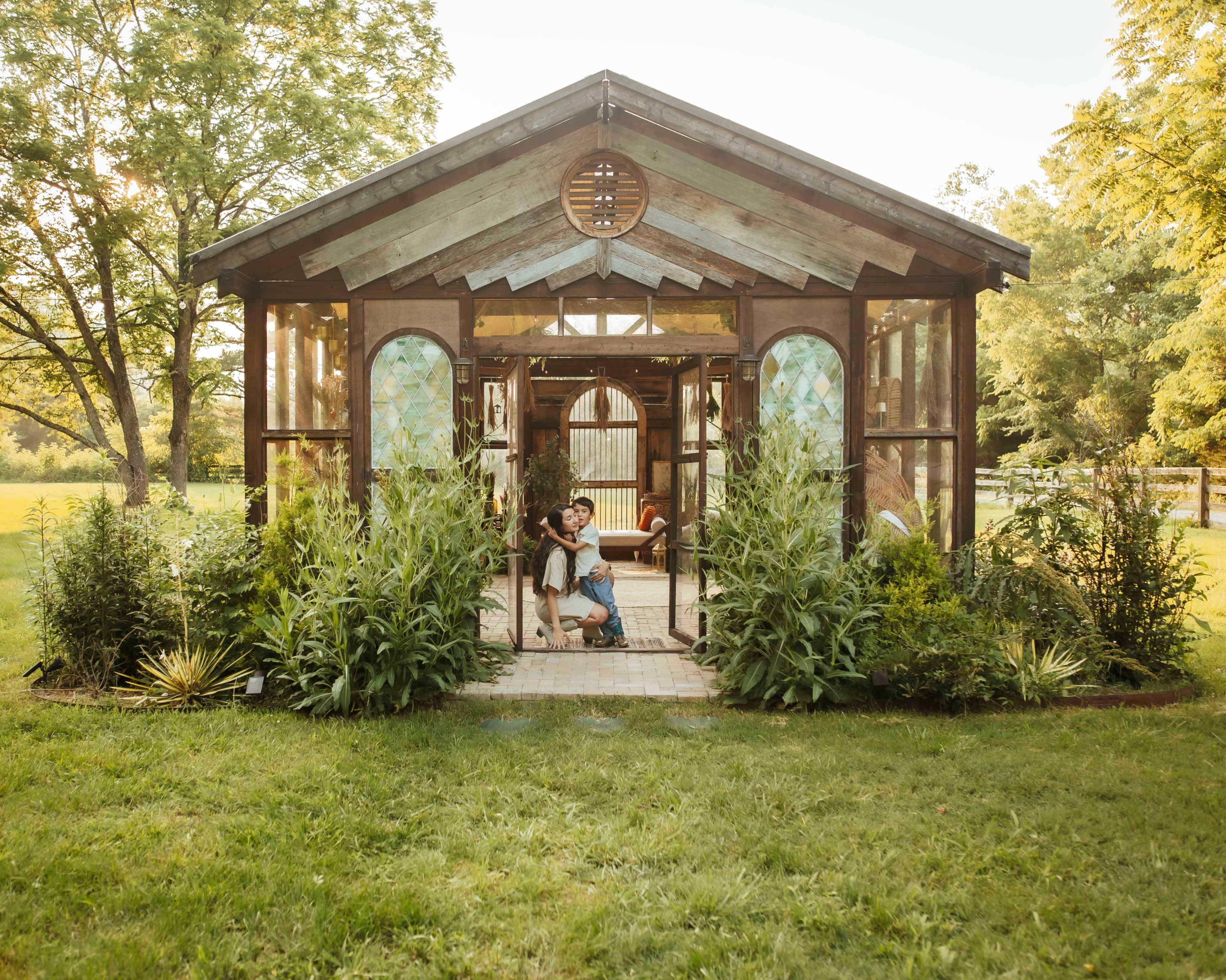 A person embraces a child in front of a wooden greenhouse surrounded by lush greenery.